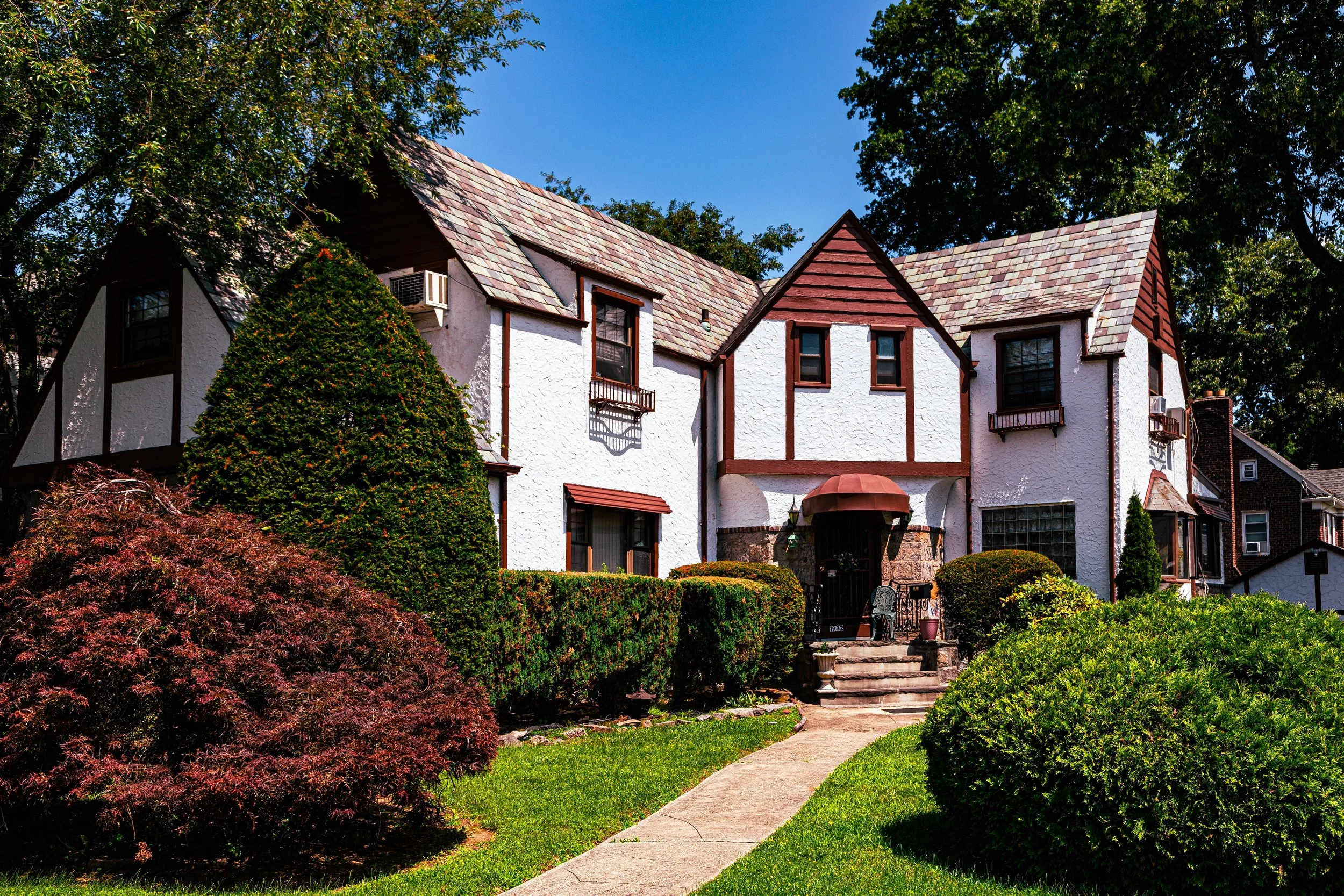A house with a Tudor-style design, white stucco walls, brown trim, and a steeply pitched shingled roof, surrounded by lush green and reddish bushes and trees.