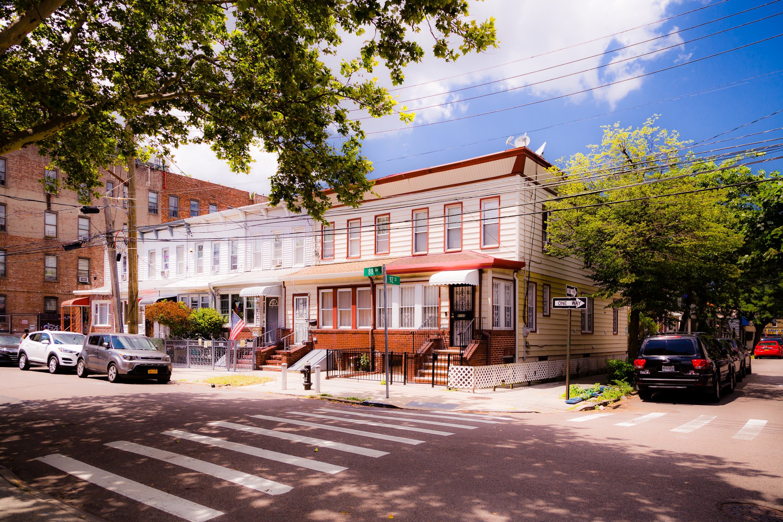 Street corner with residential buildings, parked cars, crosswalk, street signs, and trees under a partly cloudy sky.