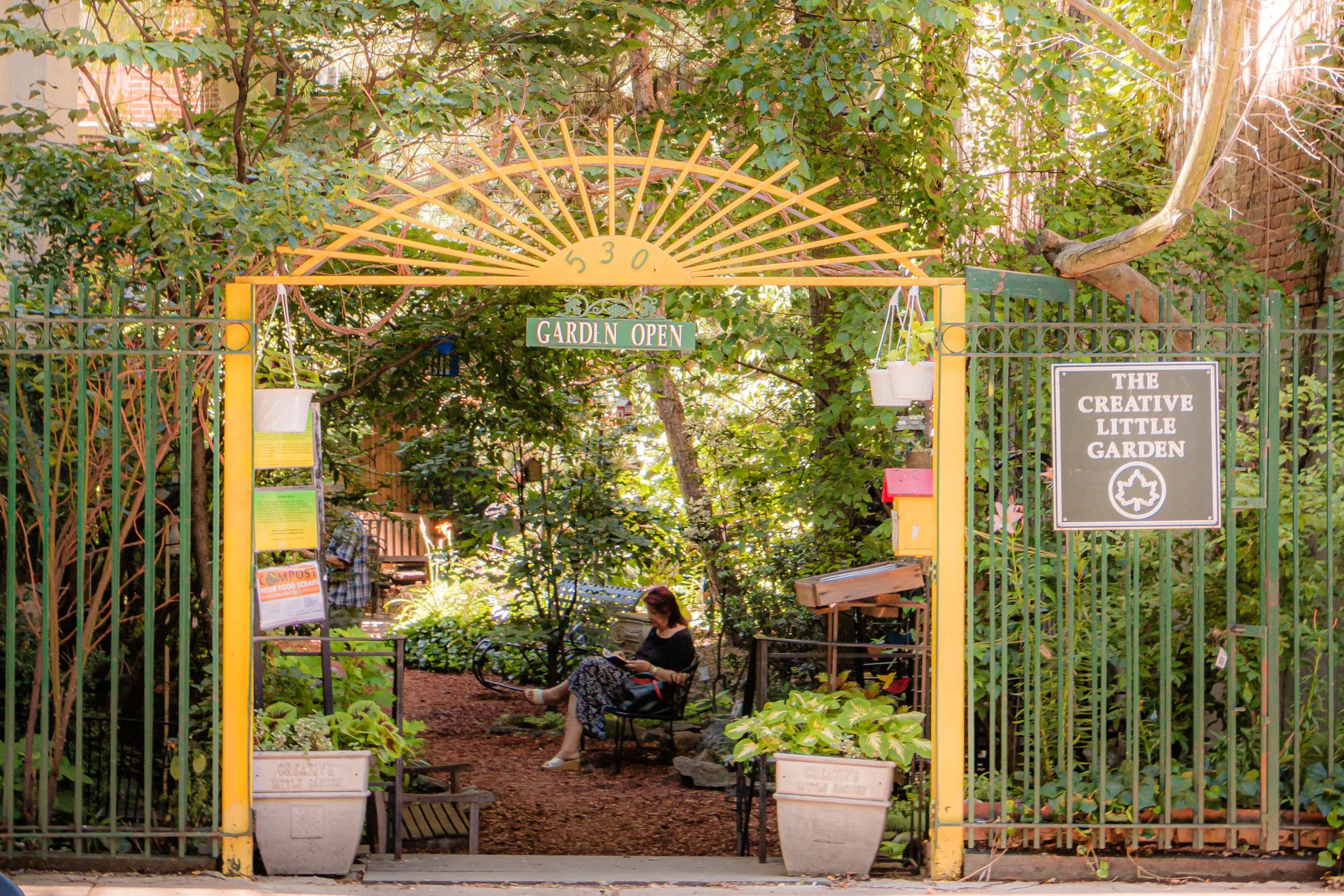 Entrance to The Creative Little Garden with a sign, gate, and a person sitting on a bench reading a book inside the garden, surrounded by lush green plants and trees.