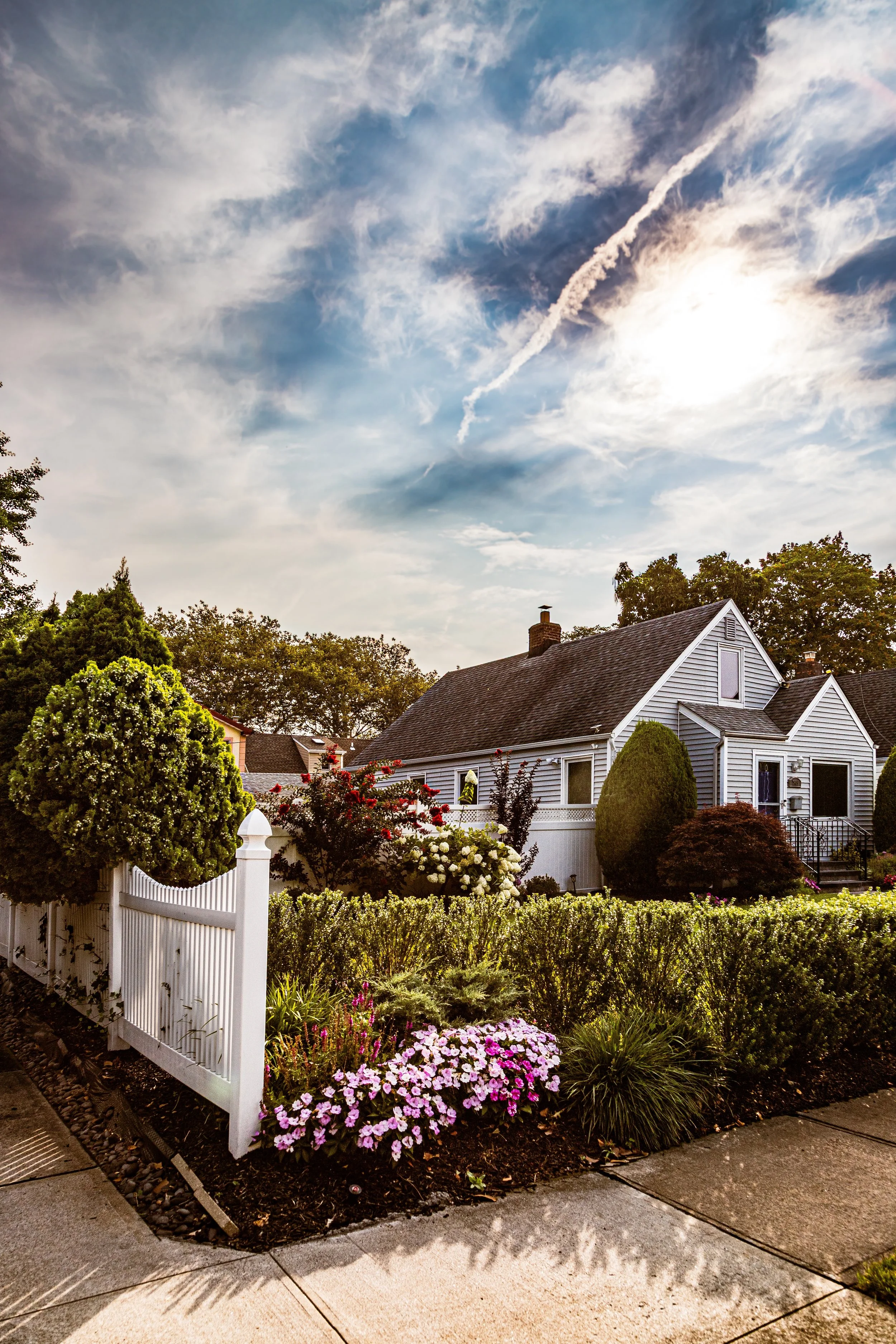 A sunny suburban house with a white picket fence and a well-maintained garden featuring pink, white, and red flowers, and trees under a partly cloudy sky.