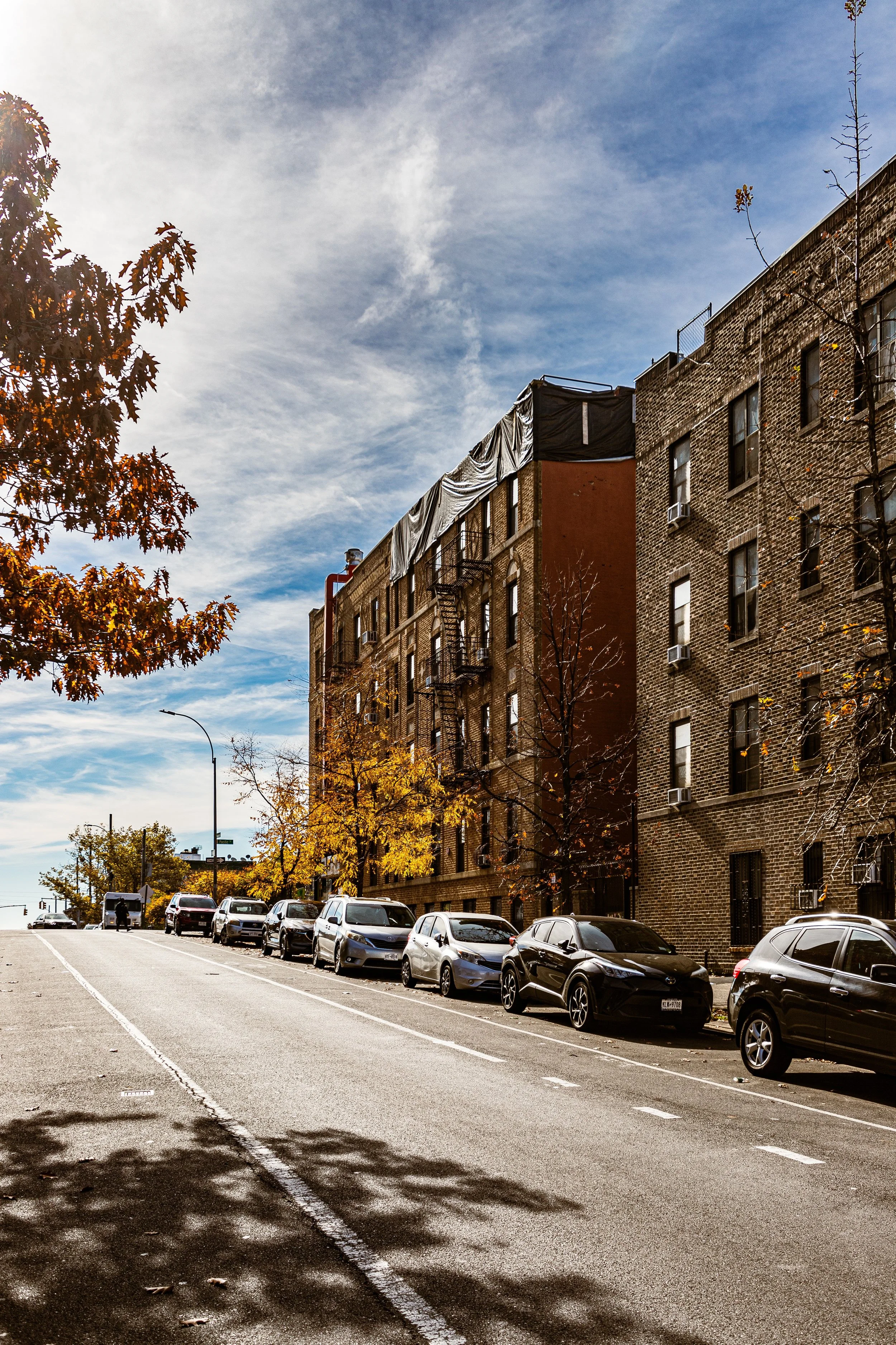 Street with parked cars, brick building, trees with autumn leaves, blue sky with clouds.