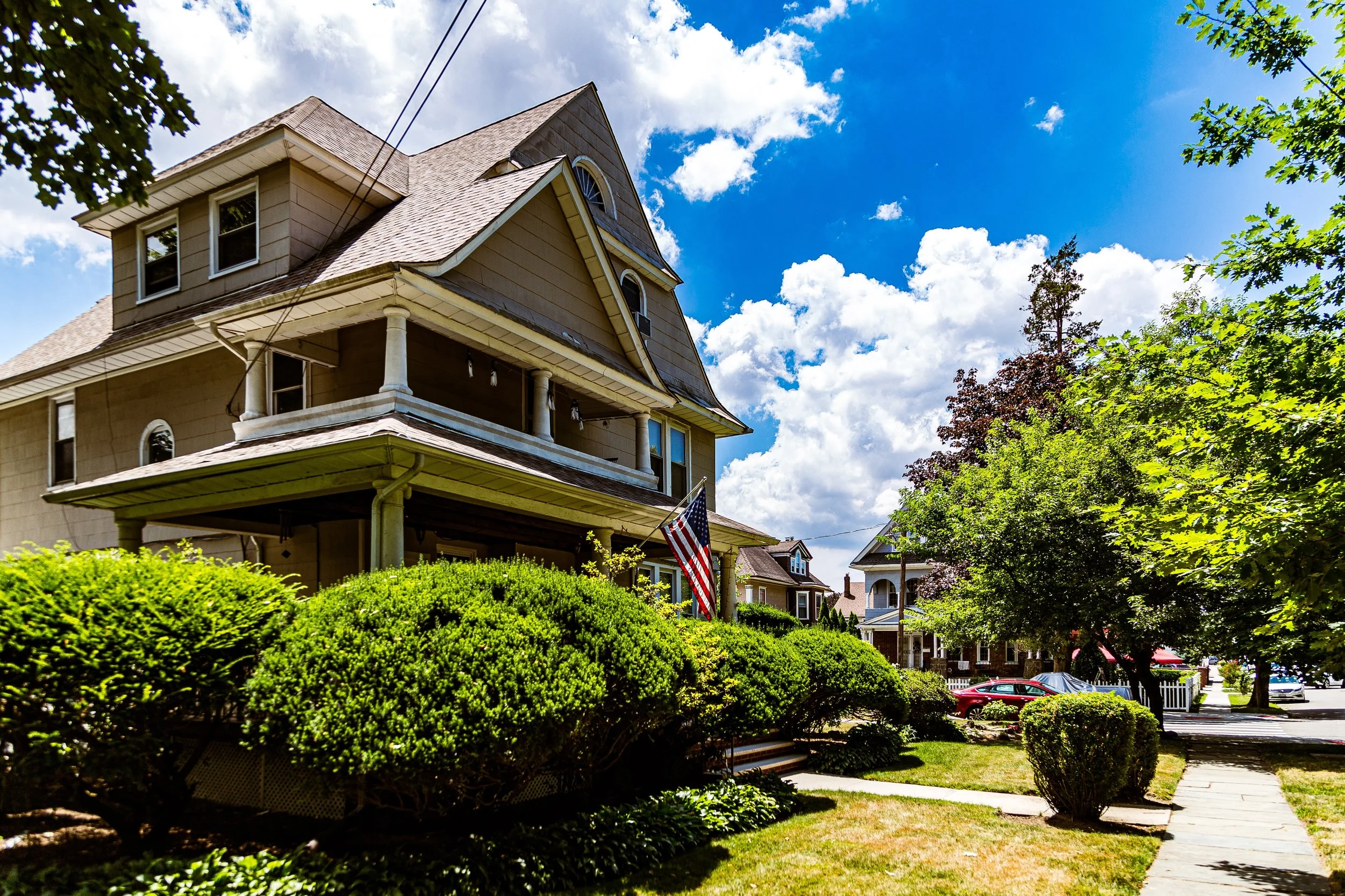 A large, multi-story house with beige siding, a gabled roof, and a front porch supported by columns. An American flag hangs from the porch. Well-maintained shrubs and trees line the sidewalk, with a bright blue sky and scattered clouds overhead.
