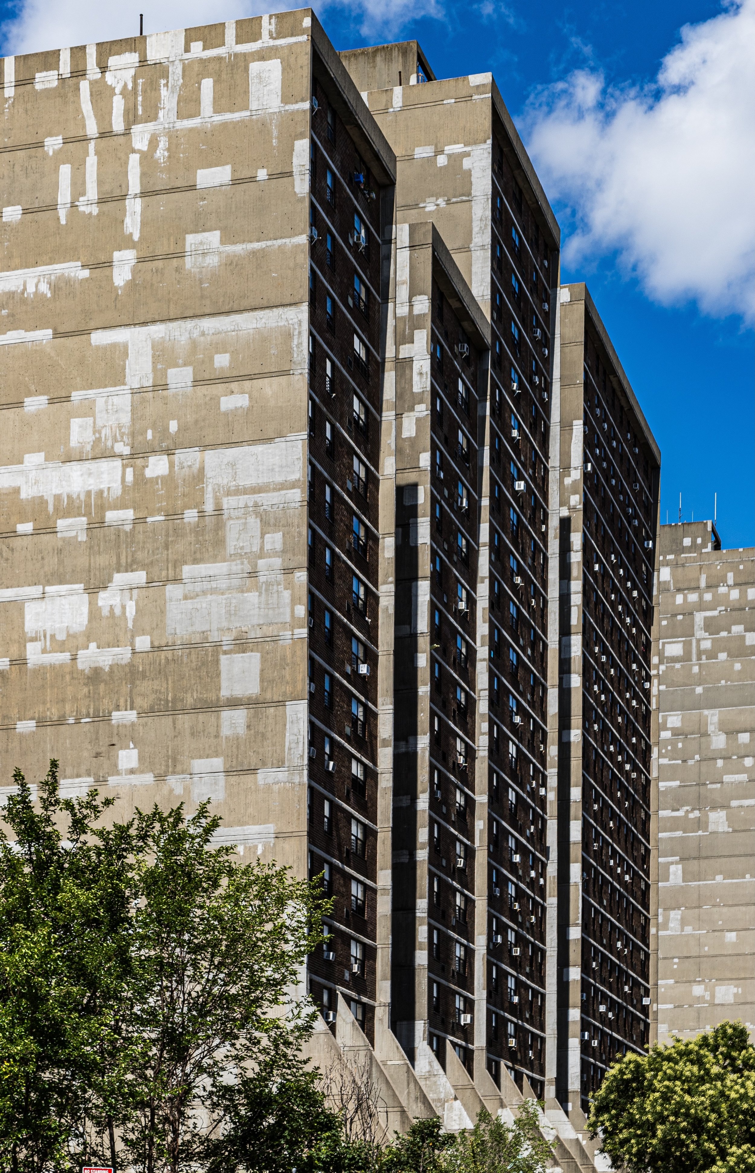 A tall, weathered concrete apartment building with dark windows and air conditioning units, set against a bright blue sky with some white clouds. Green trees are visible at the bottom of the image.