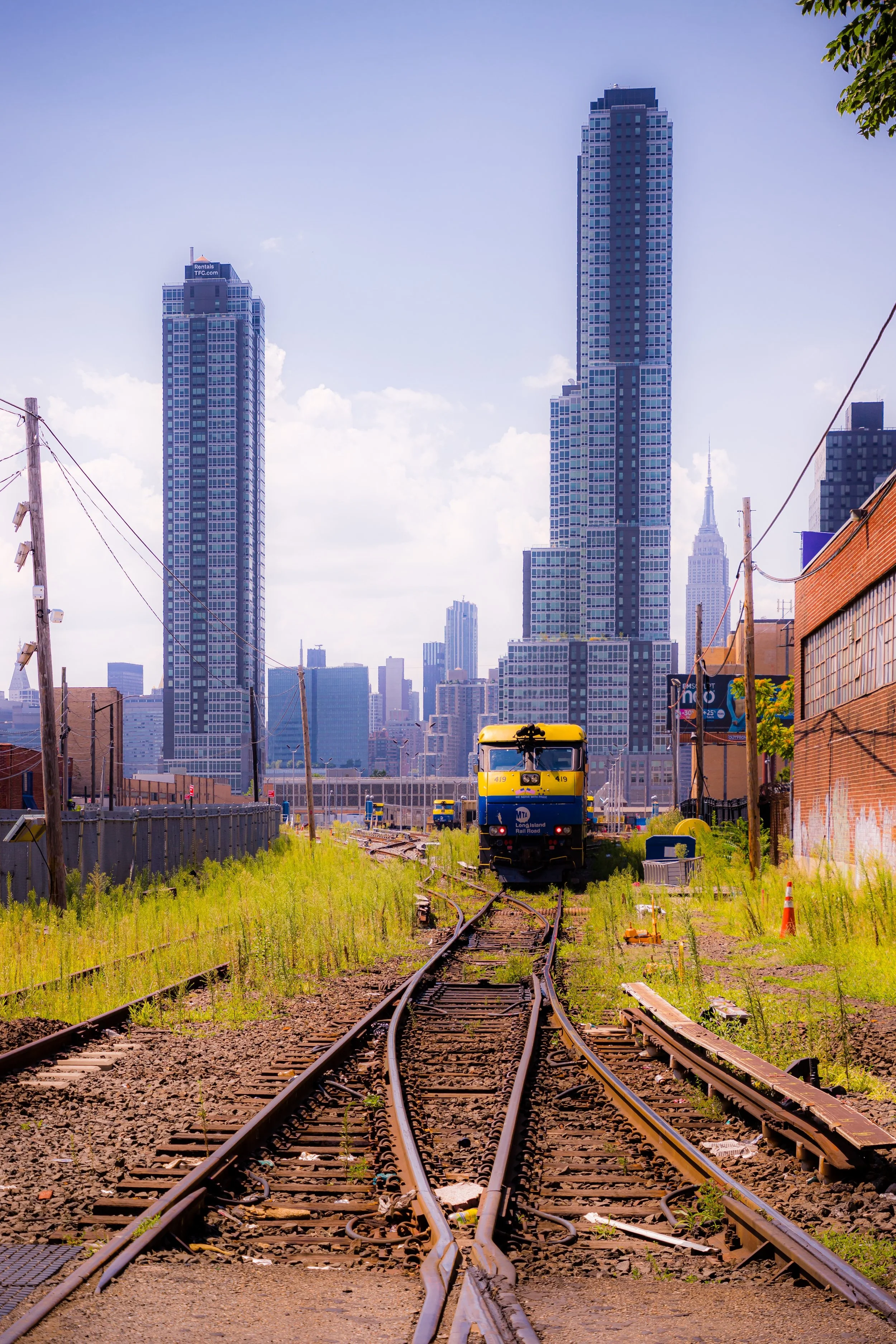 A train on railway tracks in an urban area with tall skyscrapers in the background, under a partly cloudy sky.