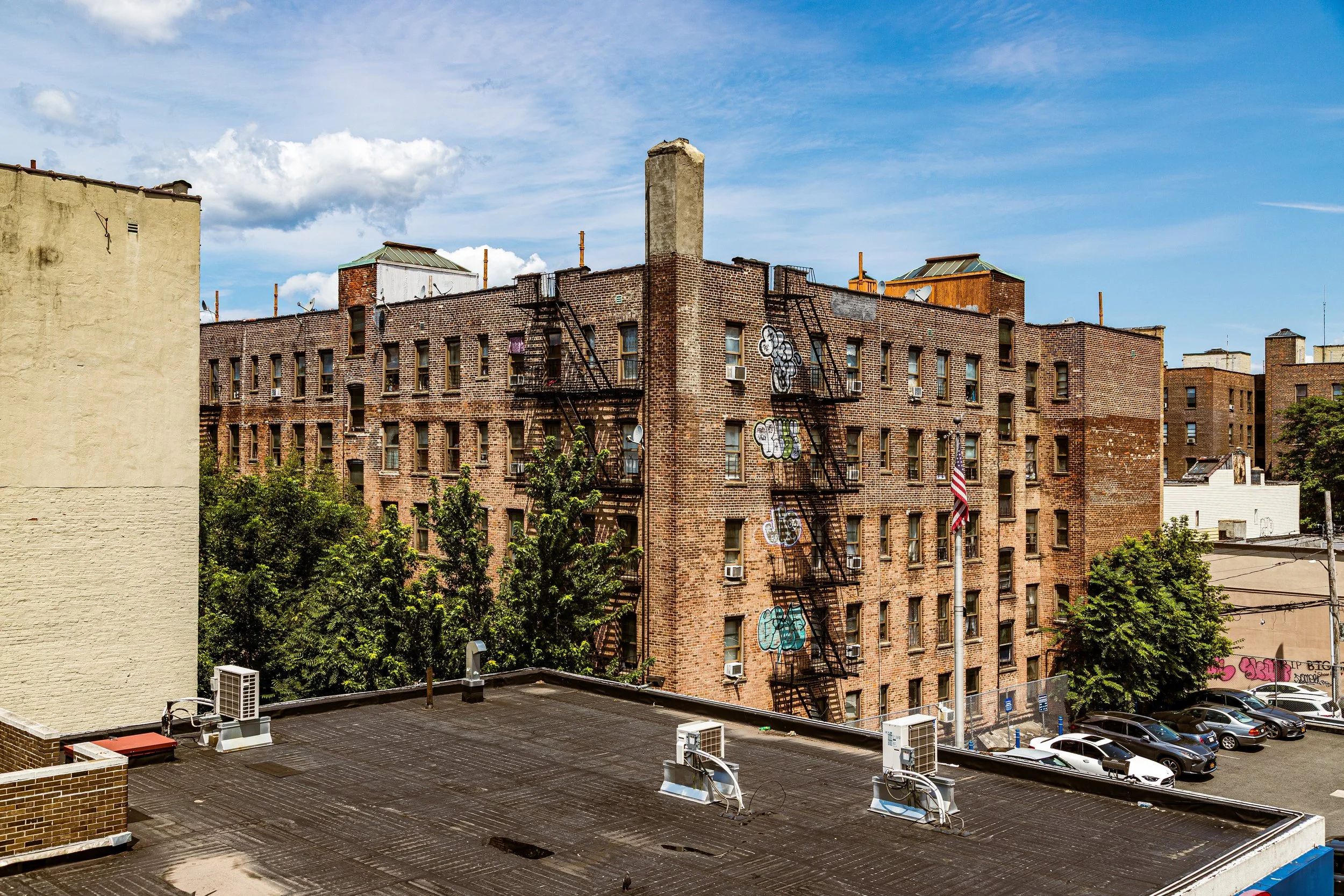 A city view showing a brick apartment building with fire escape stairs covered in graffiti, surrounded by trees, with cars parked below and a blue sky with some clouds.