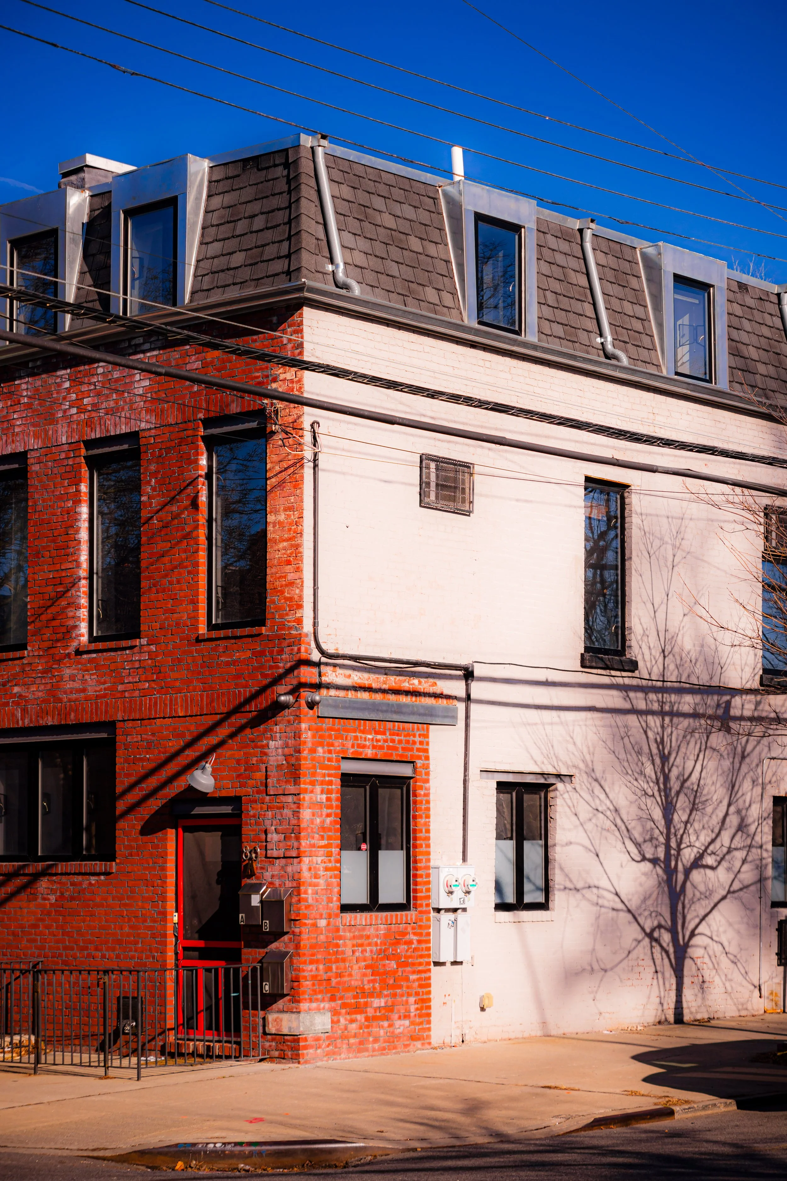 A multi-floor building with red brick and white brick exteriors, black window frames, and a dark gray shingled roof. Power lines run across the sky, and a leafless tree casts a shadow on the white wall. There is a small fence and utility meters near the front door.