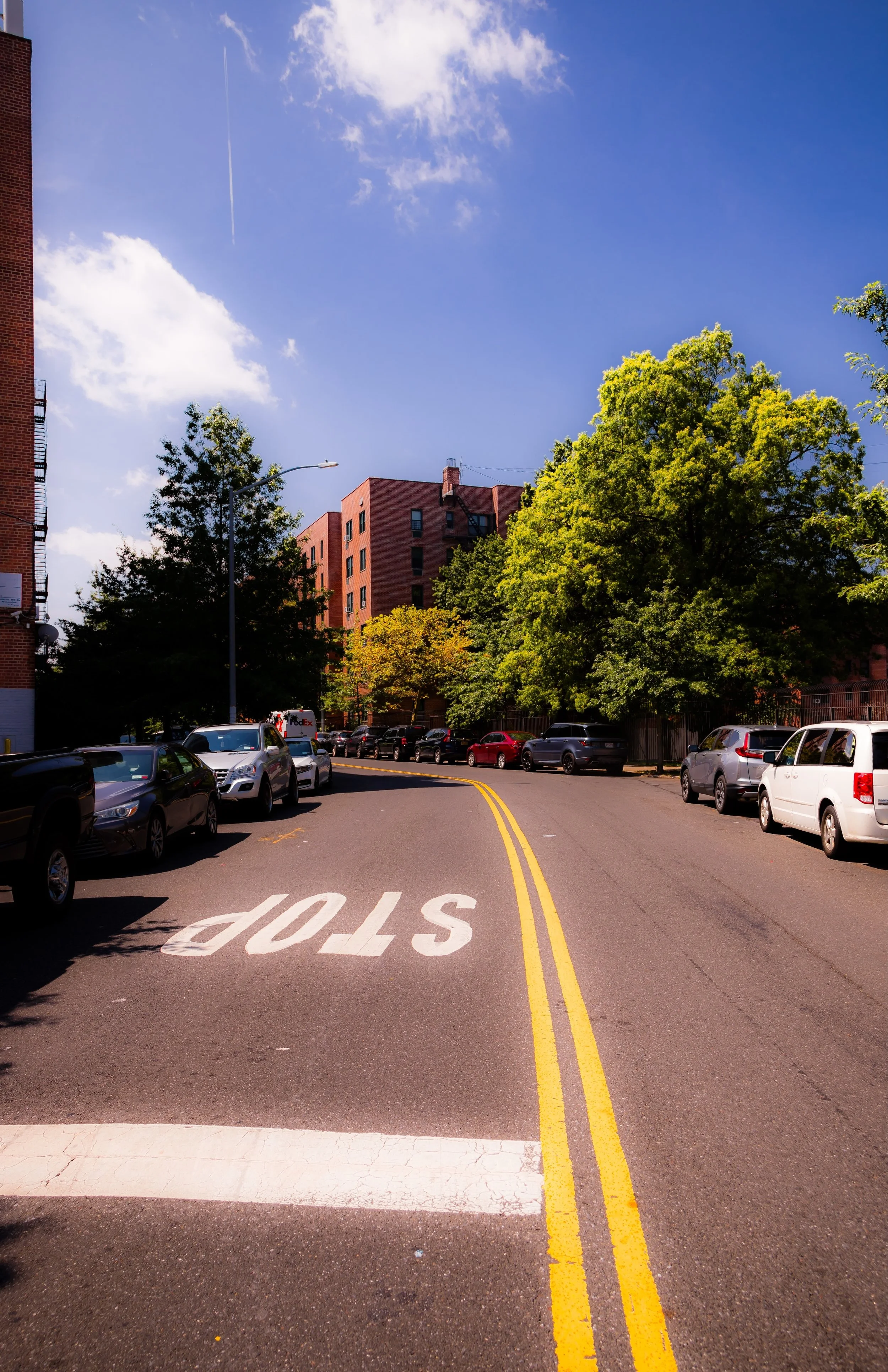 City street with parked cars, trees, apartment buildings, and a blue sky with clouds and a contrail