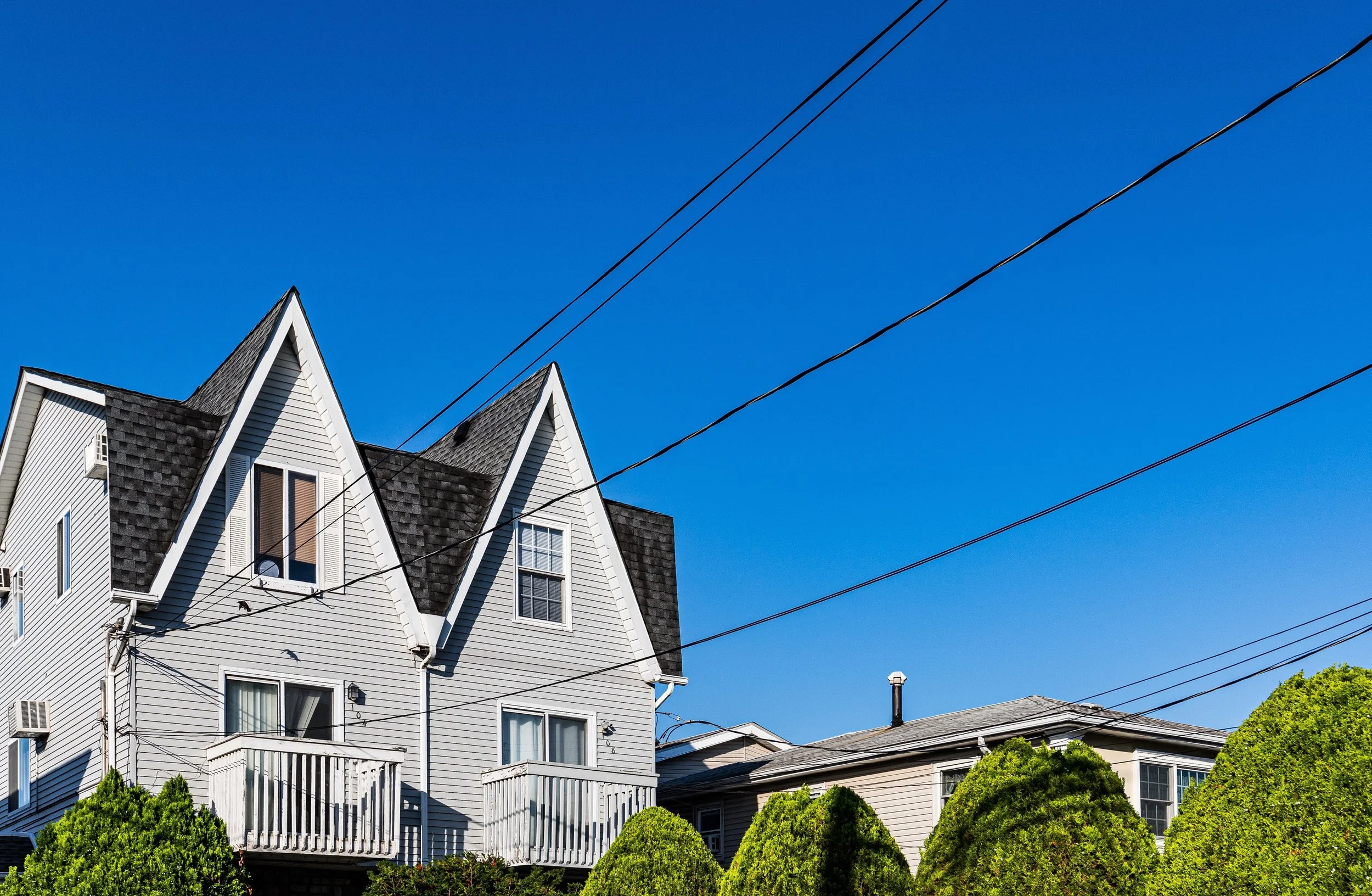 A row of residential houses under a clear blue sky, with power lines overhead and green bushes in the foreground.