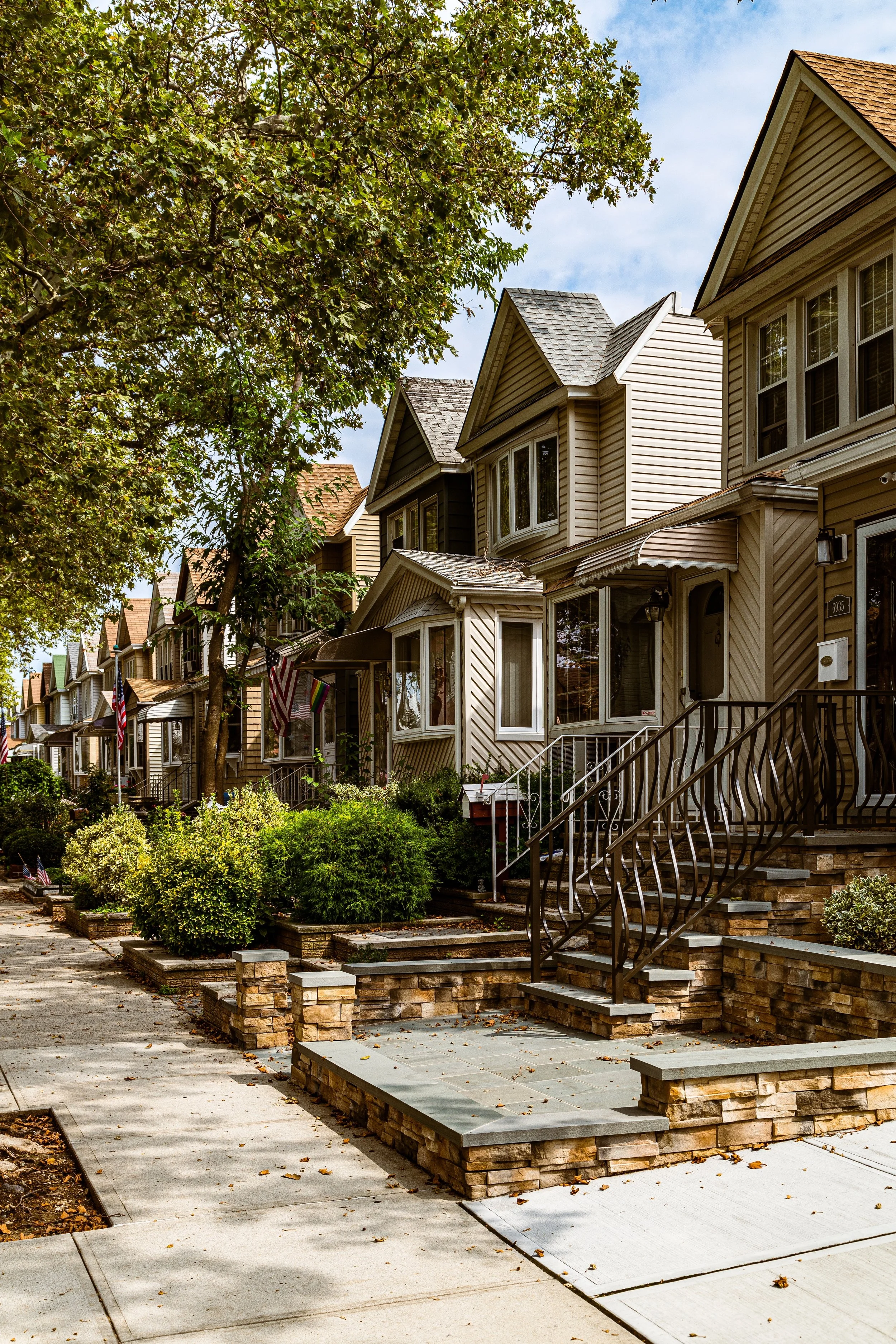 Row of beige and brown houses with small front yards and stairs, trees along the sidewalk, and blue sky with clouds overhead.