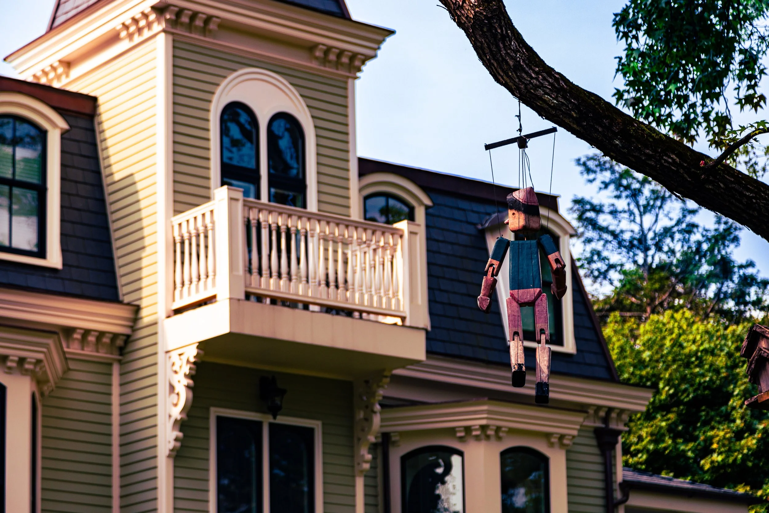 A decorative wooden puppet hanging from a tree branch in front of a large Victorian-style house with beige siding, black roof, and arched windows.