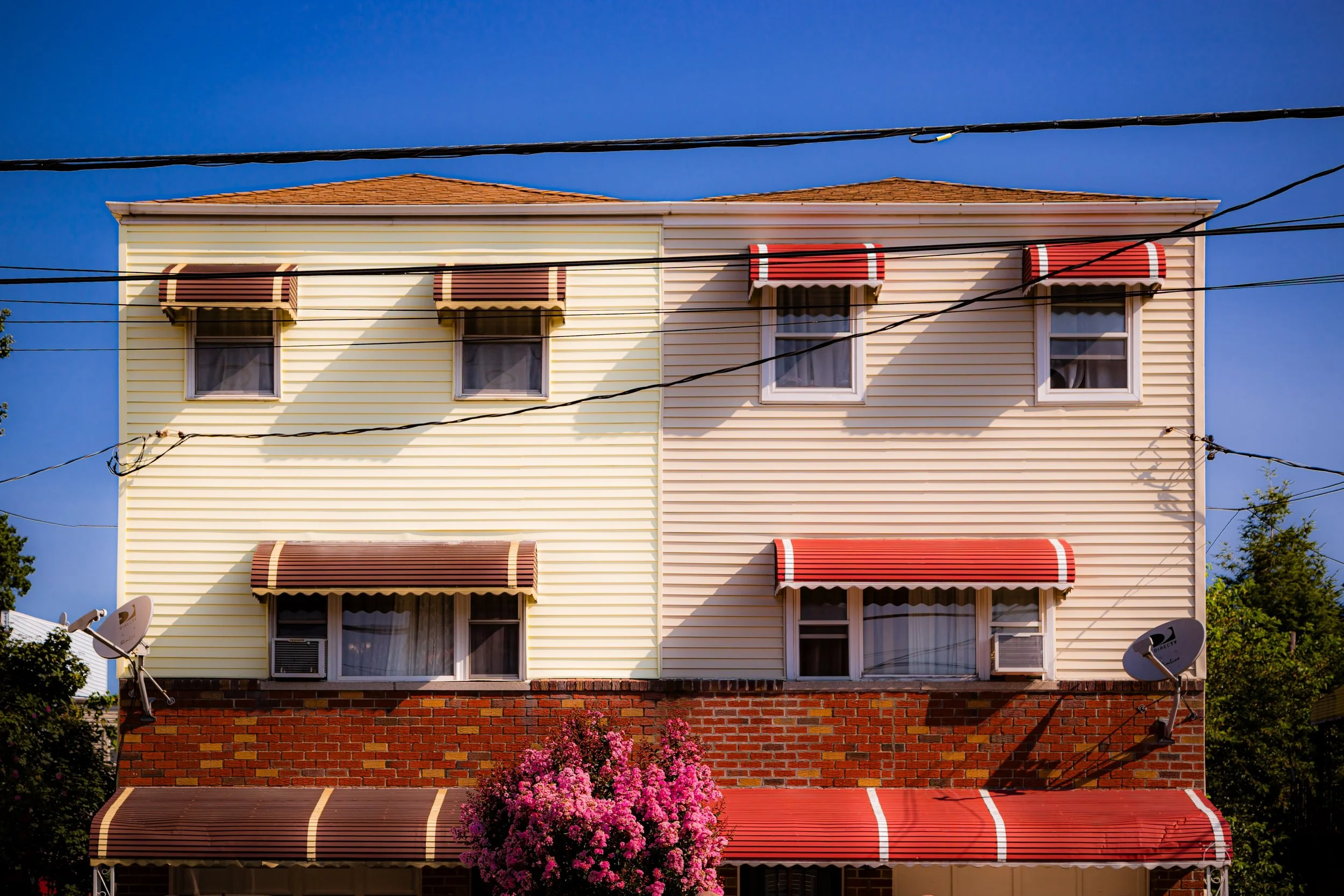 A three-story residential building with yellow and beige siding, red and brown striped awnings on the windows, brick lower facade, and a vibrant pink flowering tree in front. Overhead power lines are visible against a bright blue sky.