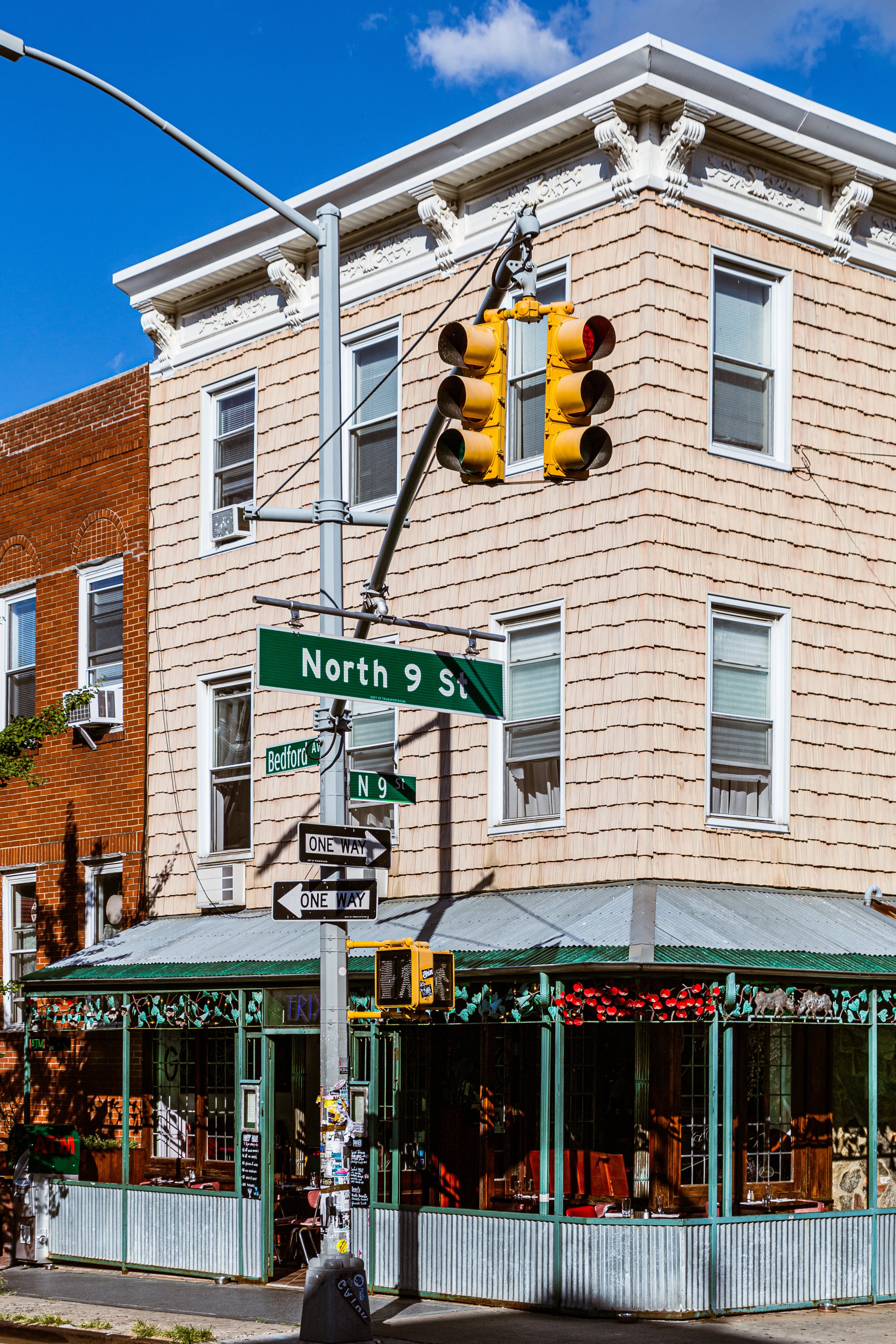 Street corner with traffic light, street signs for North 9th Street and Bedford Avenue, a one-way sign, a green and glass enclosed restaurant patio, and a beige building with white decorative crown molding under a blue sky.