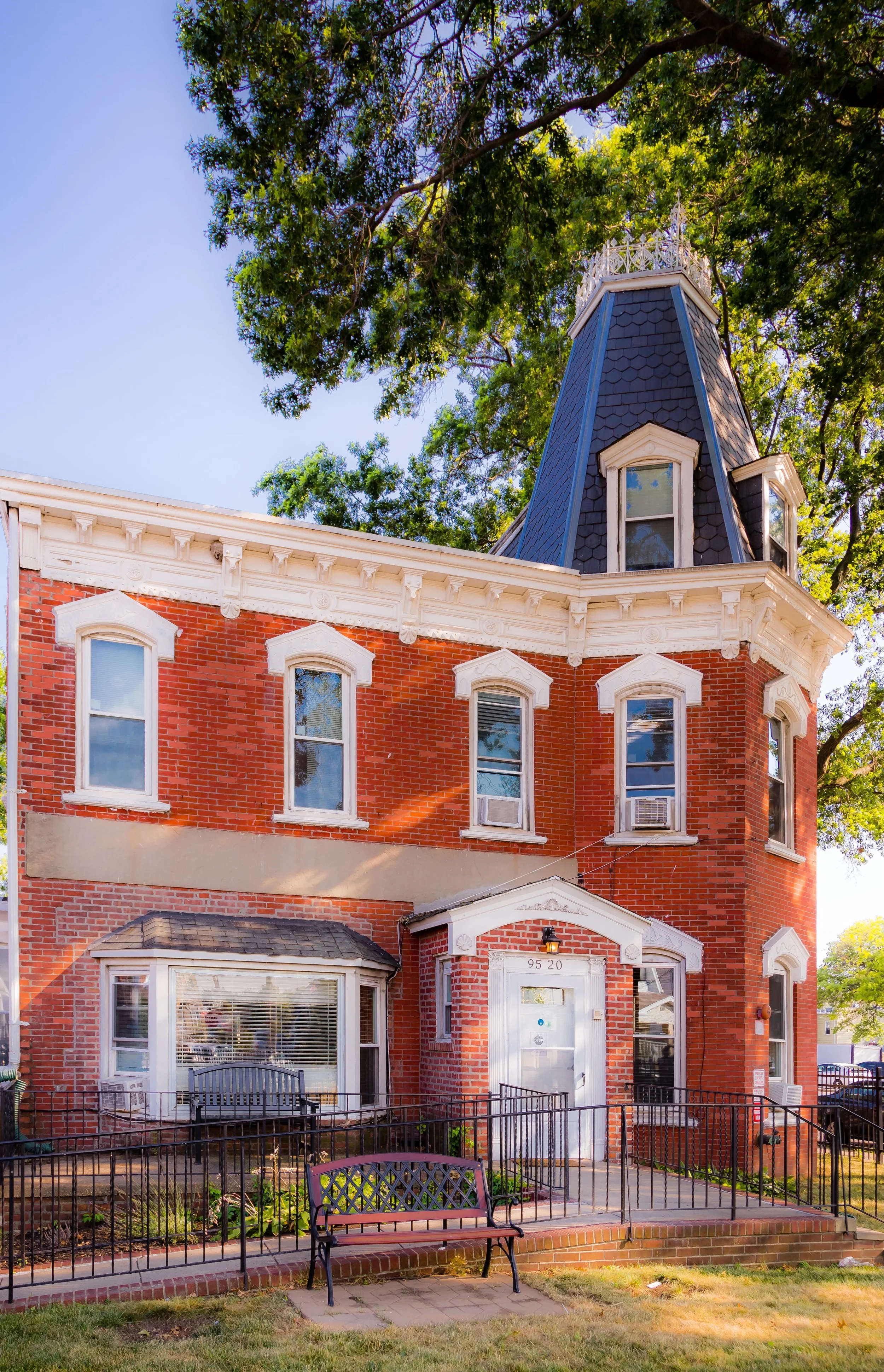 A three-story red brick house with white trim, multiple windows with white frames, and a rounded bay window on the left side. The house features a distinctive tower with a pointed dark shingle roof and a white decorative railing at the top. A small porch with a white door and house number 9520 is accessible via a short set of steps, enclosed by a black metal fence. A pink bench is placed in front of the house, with a flower bed and a tree with green foliage in the background.