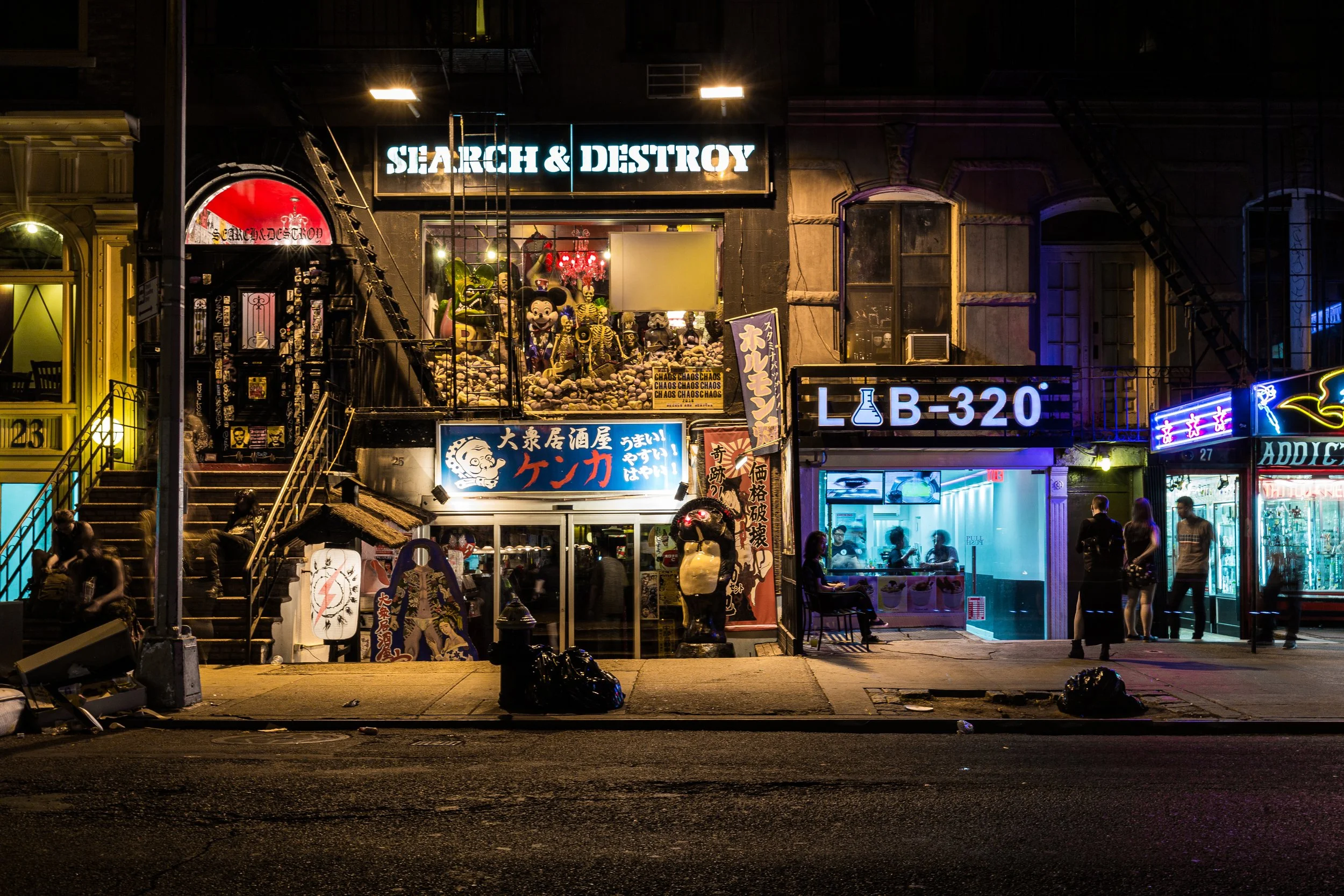 Night scene of a city street with illuminated storefronts, including a bar or restaurant with a sign that reads 'SEARCH & DESTROY,' and other businesses with neon signs and people outside.