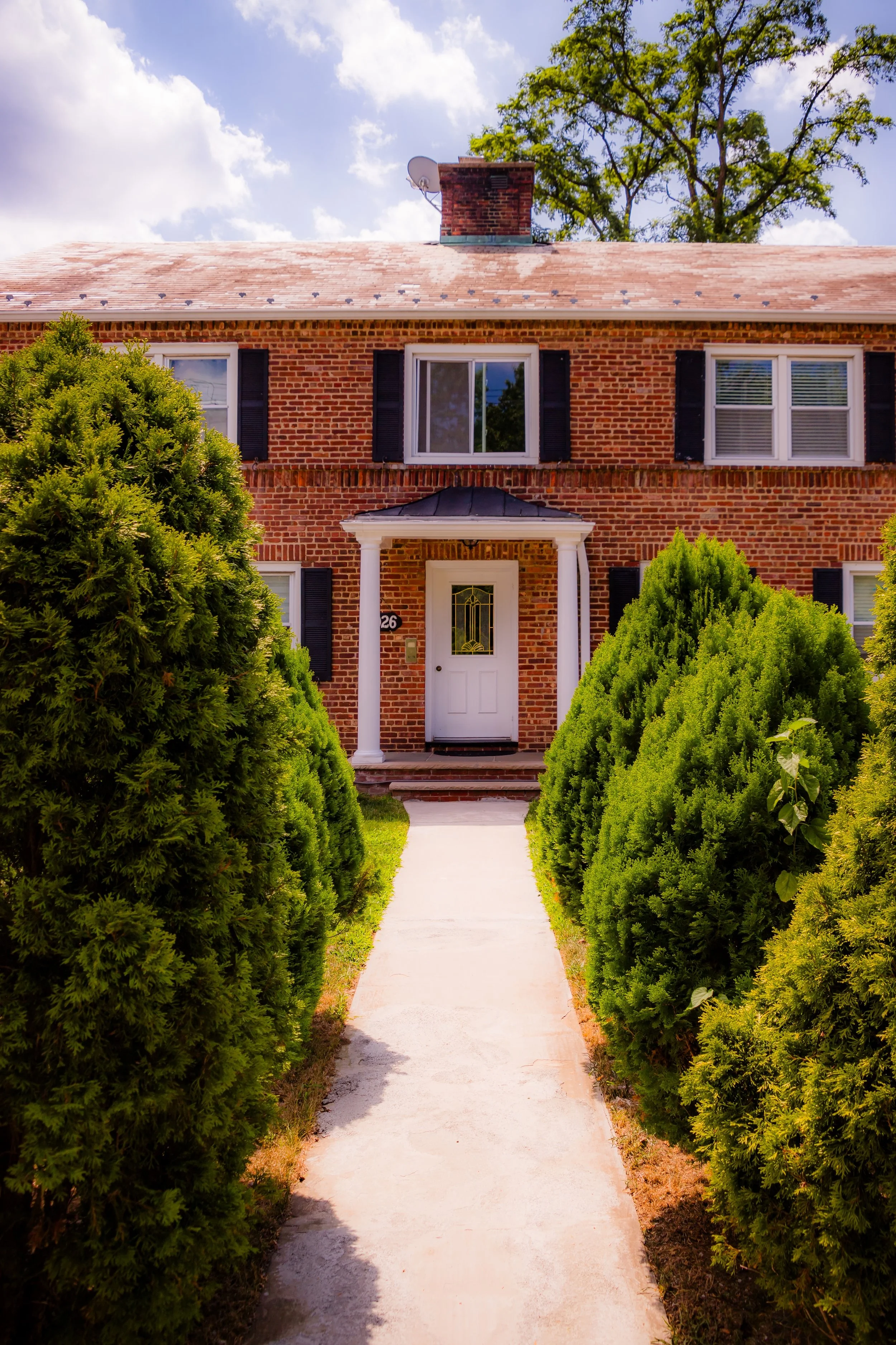 Brick house with front porch, white door, black shutters, and lush green bushes along sidewalk.