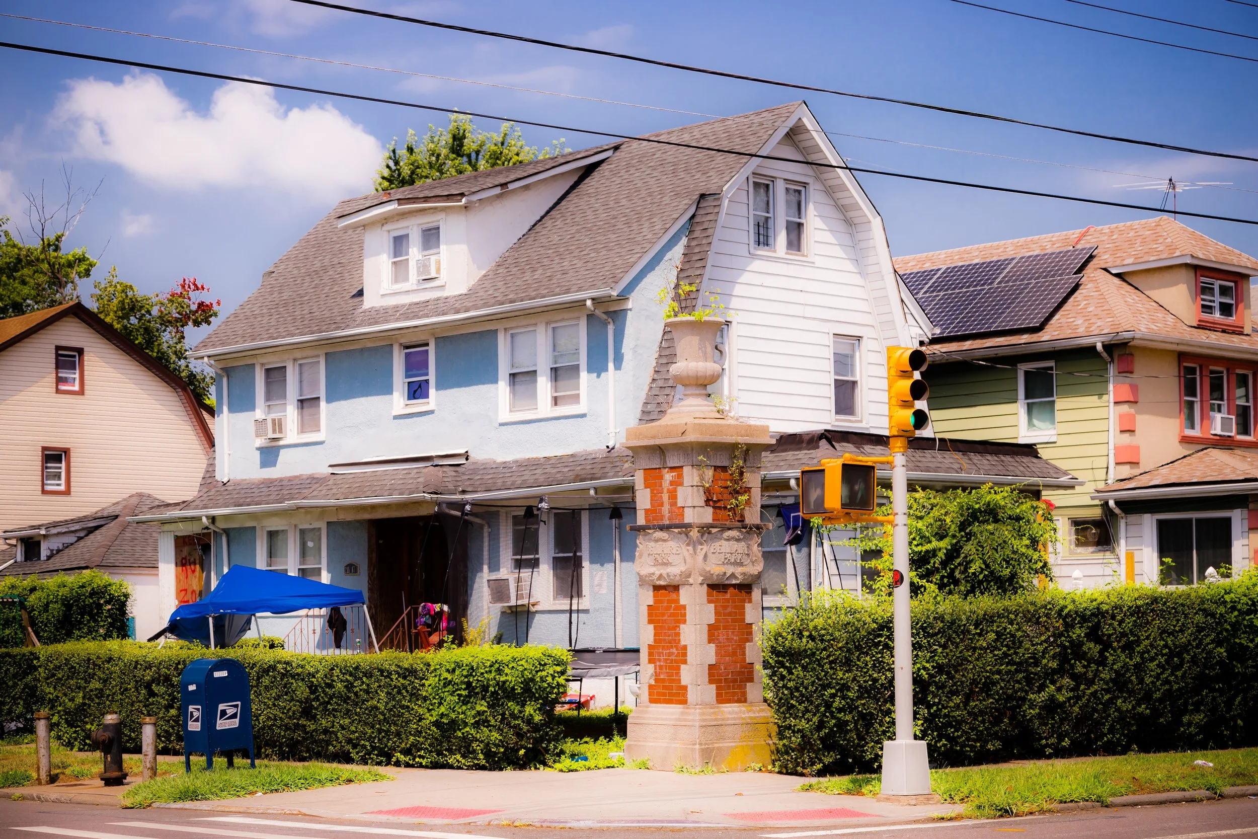 Older two-story house with blue and white siding, a steep gable roof, and a small dormer window. In front, there's a brick monument, greenery, a blue mailbox, and a traffic light set against a blue sky with some clouds.