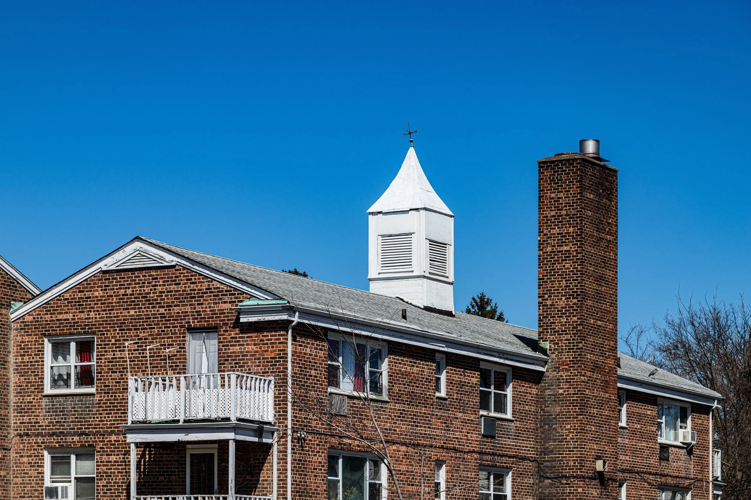 Brick apartment building with white balconies, a tall brick chimney, and a white steeple with a weather vane against a clear blue sky.