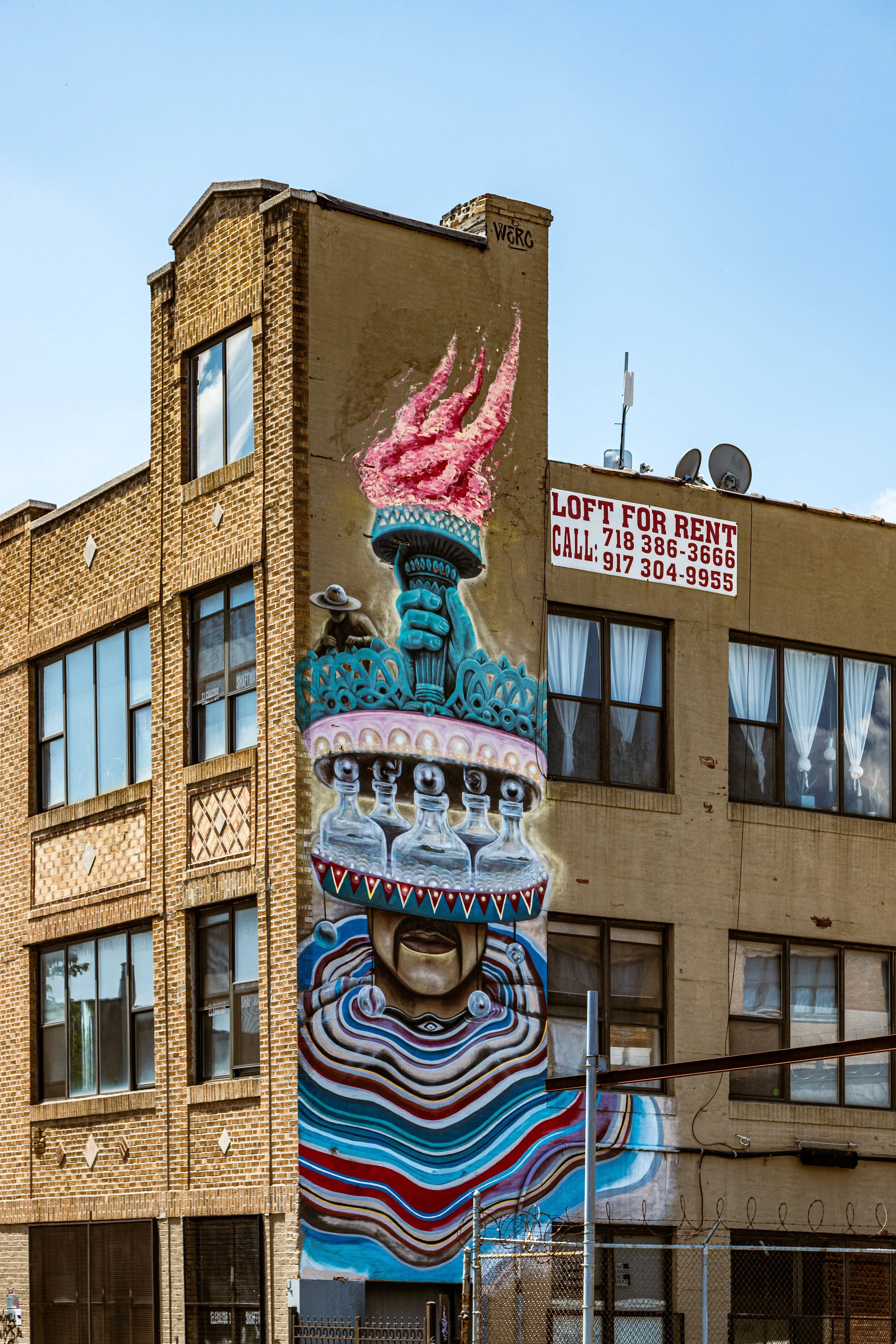 Colorful mural of a woman with flowing, striped hair, wearing large earrings and a necklace, with a layered, decorative headpiece resembling bottles and a flaming torch on her head, painted on the side of a building.