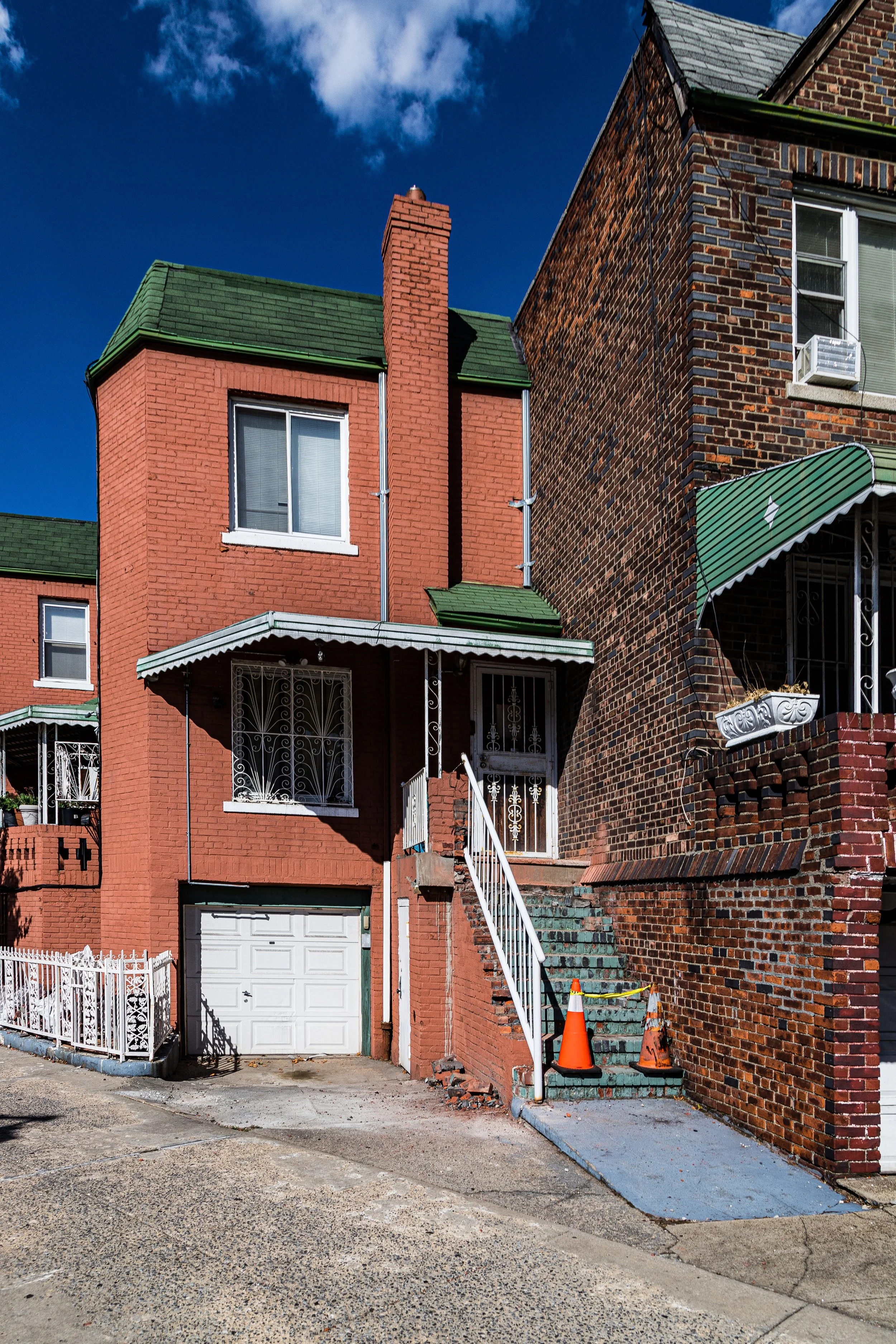 Two adjacent houses, one painted red with green roofing, and the other brick with a green awning, on a sunny day with a blue sky. There are two orange traffic cones and a yellow caution tape on the stairs leading to the red house.