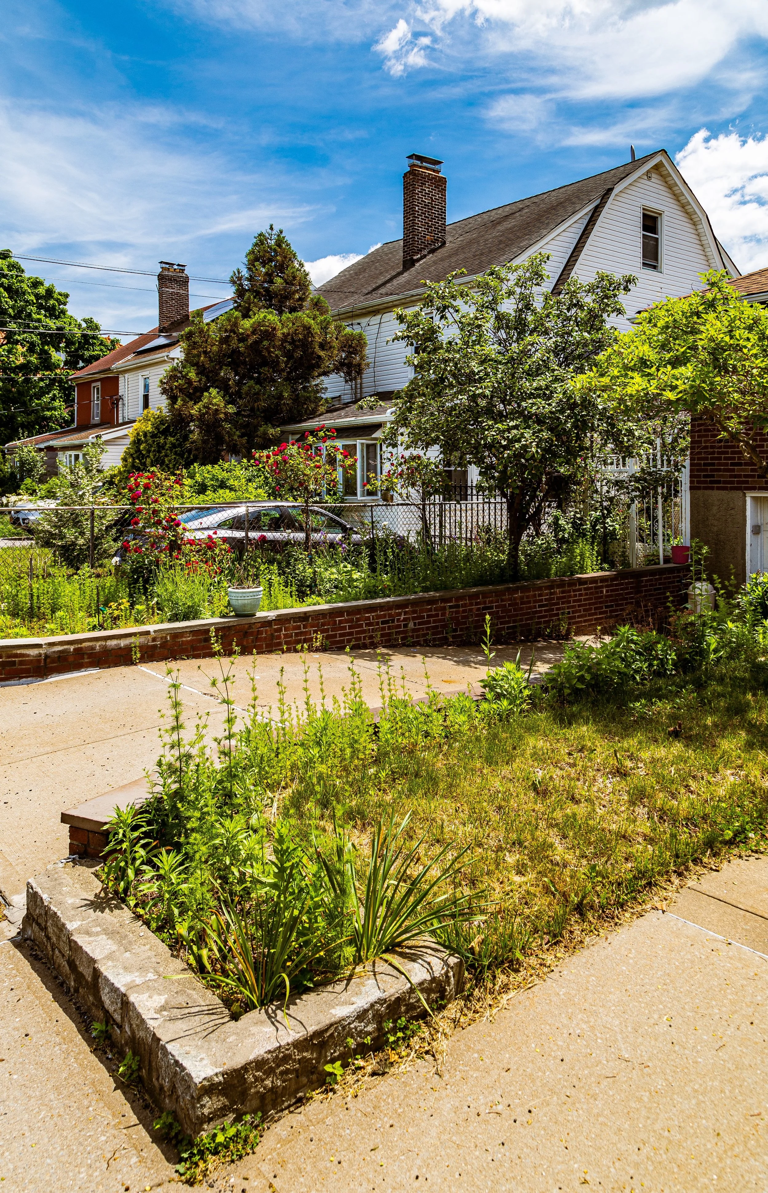 A suburban backyard with a small garden, trees, and flowering bushes in front of a white house with a steep roof and brick chimney, under a partly cloudy blue sky.