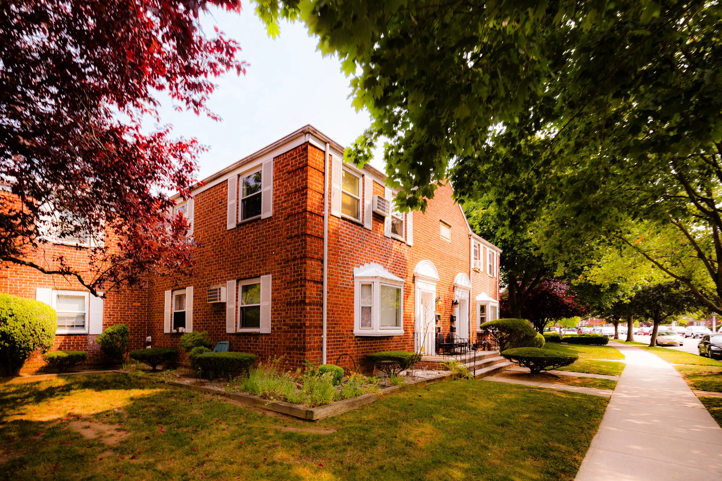 A brick apartment building with white trim and shutters, surrounded by green trees and a landscaped yard with bushes and flower beds, along a sidewalk.