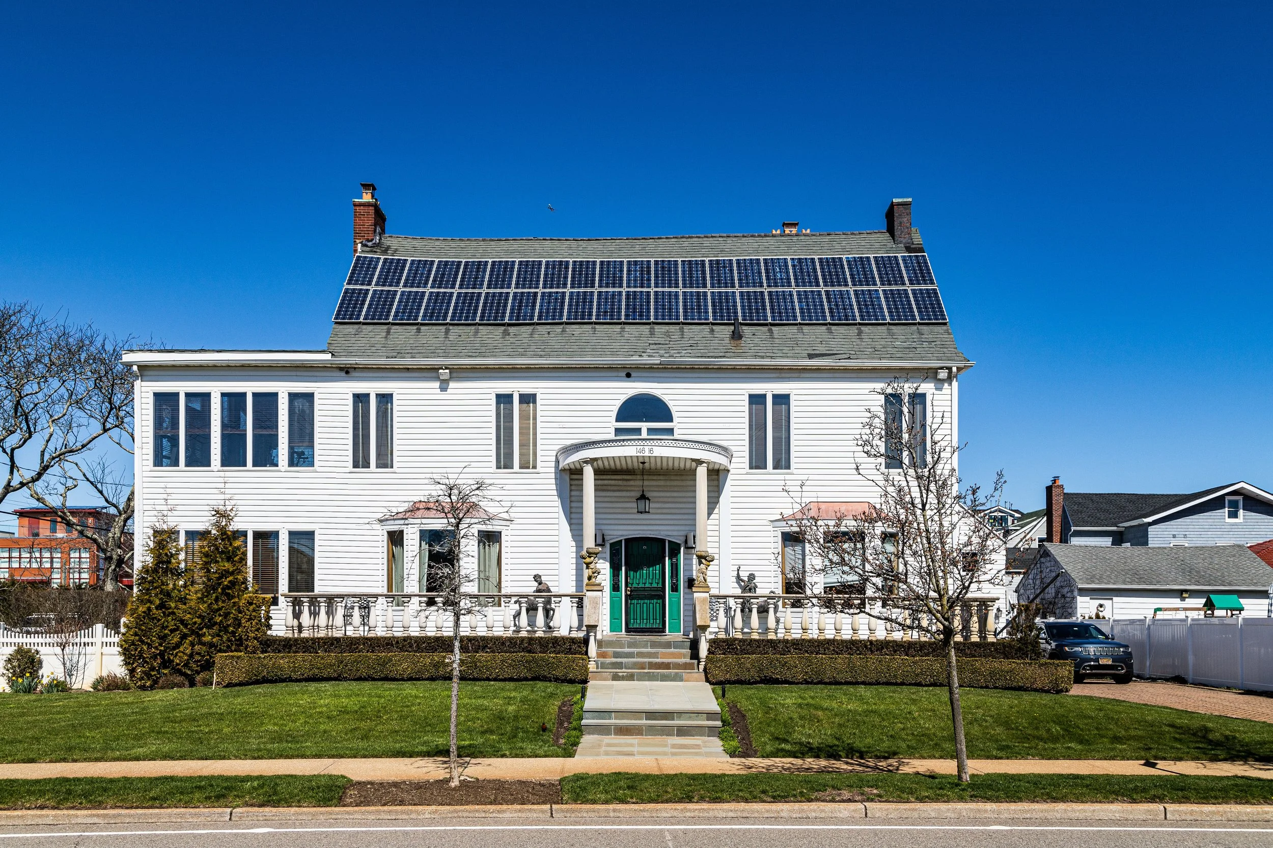 Large white house with solar panels on roof, green front door, steps leading up to porch, landscaped yard with trees, and a driveway with a black vehicle