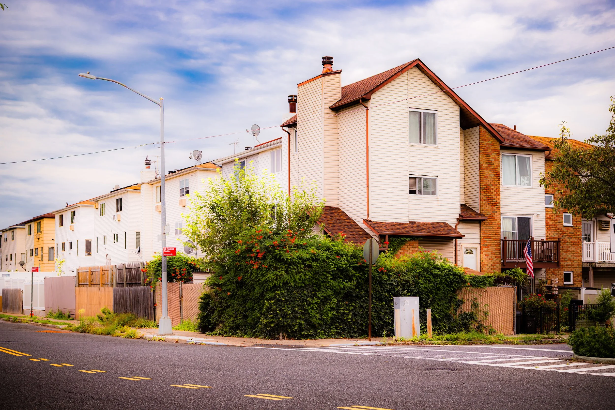 Multi-story residential buildings with brick and siding exteriors, trees, bushes, a street with double yellow lines, a streetlight, power lines, and a cloudy sky.