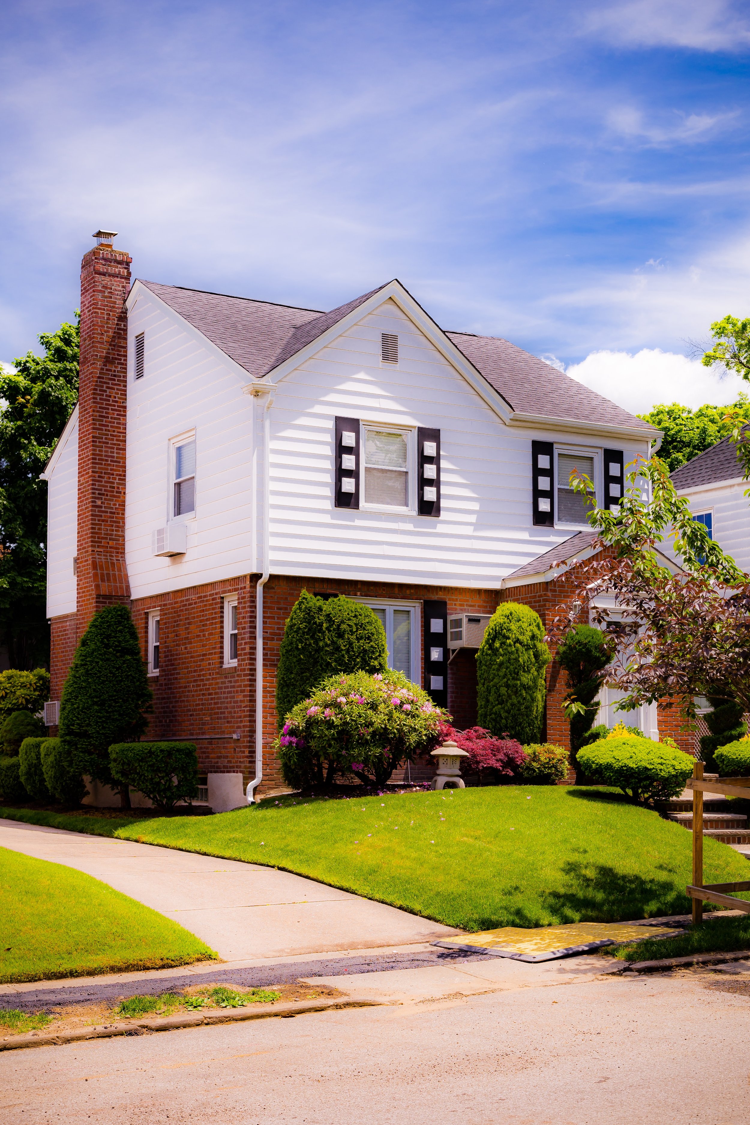 A two-story house with brick and white siding exterior, surrounded by manicured lawn and bushes, under a partly cloudy blue sky.
