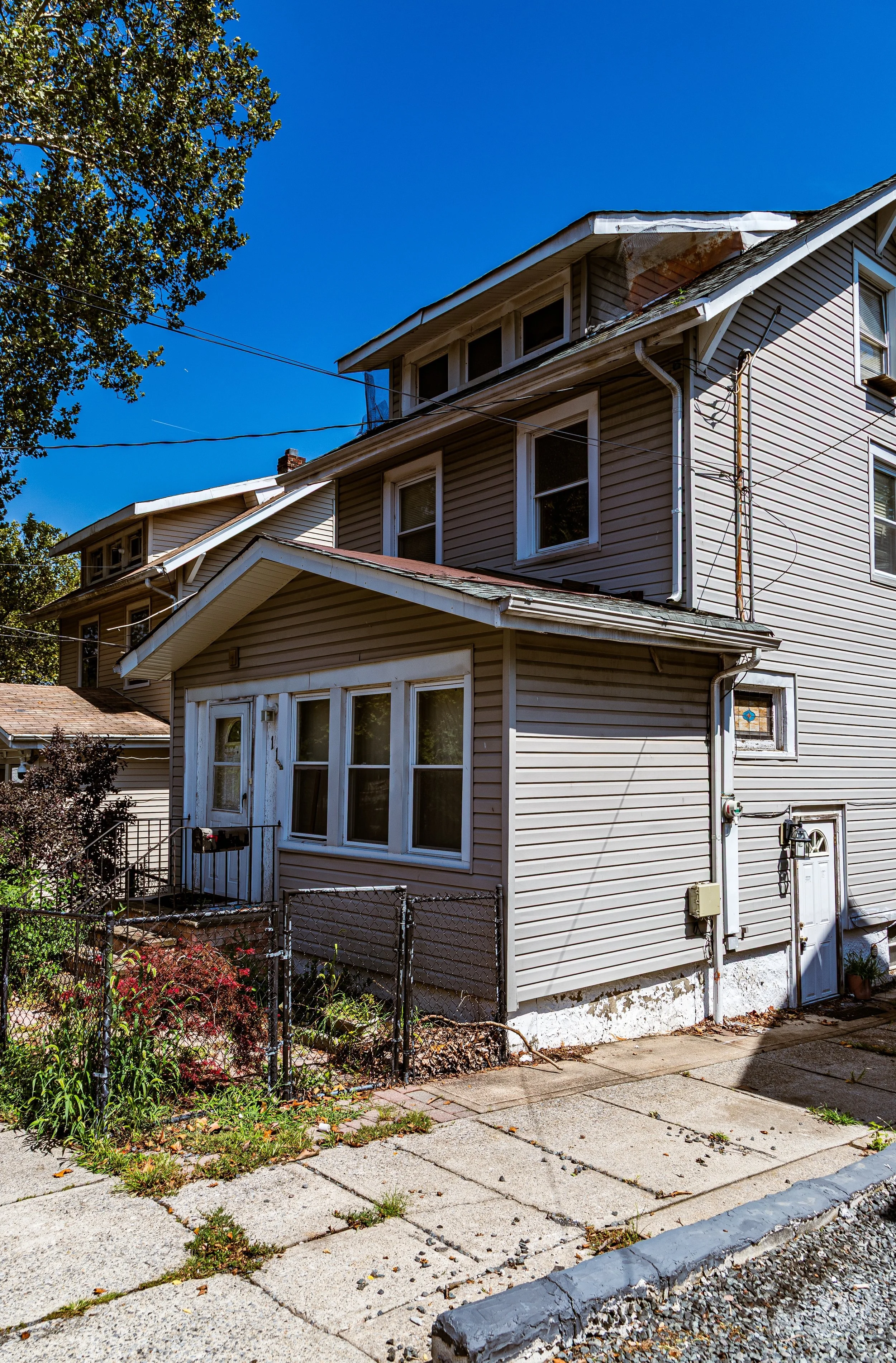 A multi-story house with beige siding, a small front yard enclosed by a black metal fence, and a concrete sidewalk in front under a clear blue sky.
