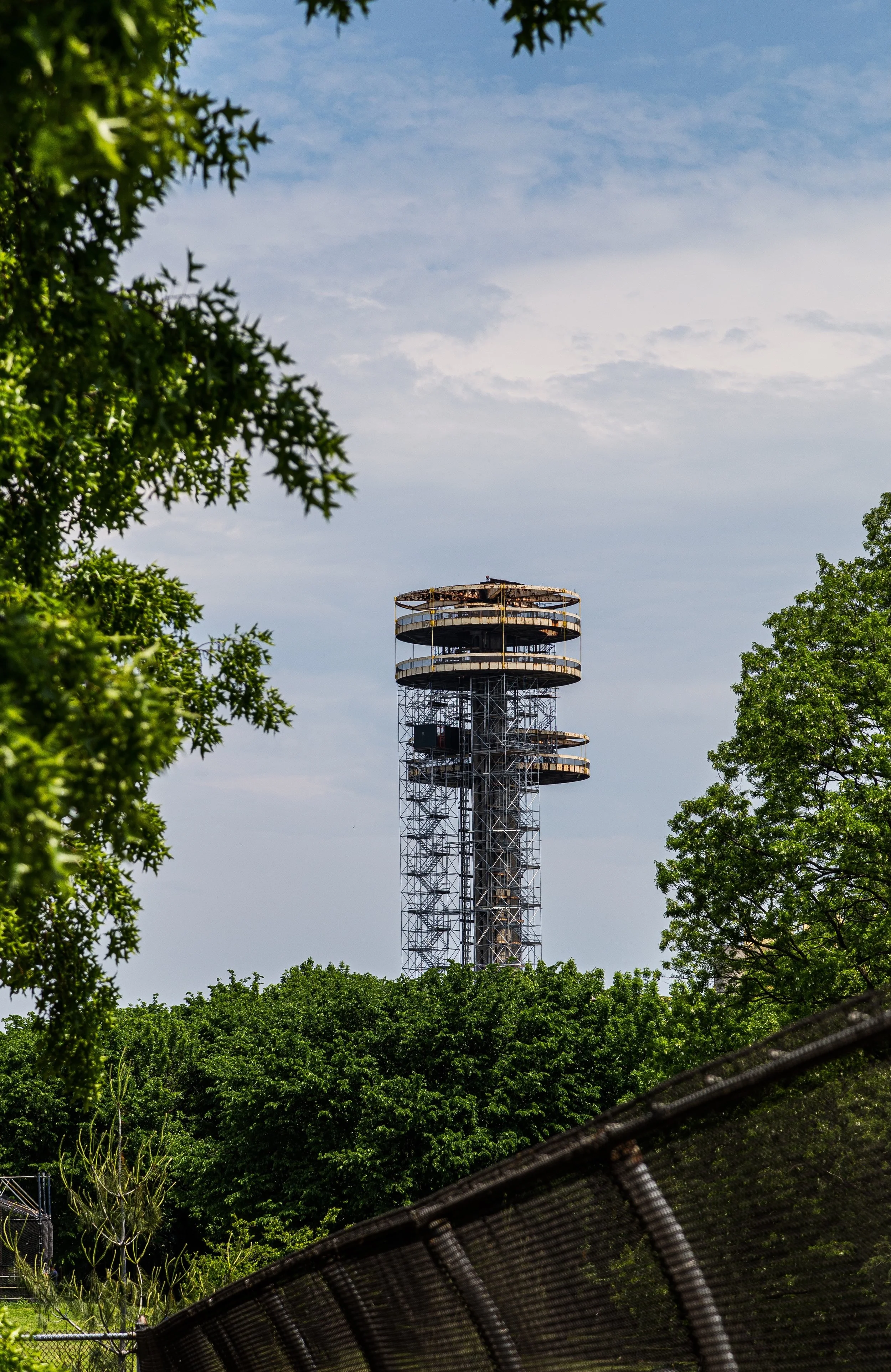 A tall observation tower with a spiral staircase around the exterior, visible above a dense canopy of trees, under a partly cloudy sky.