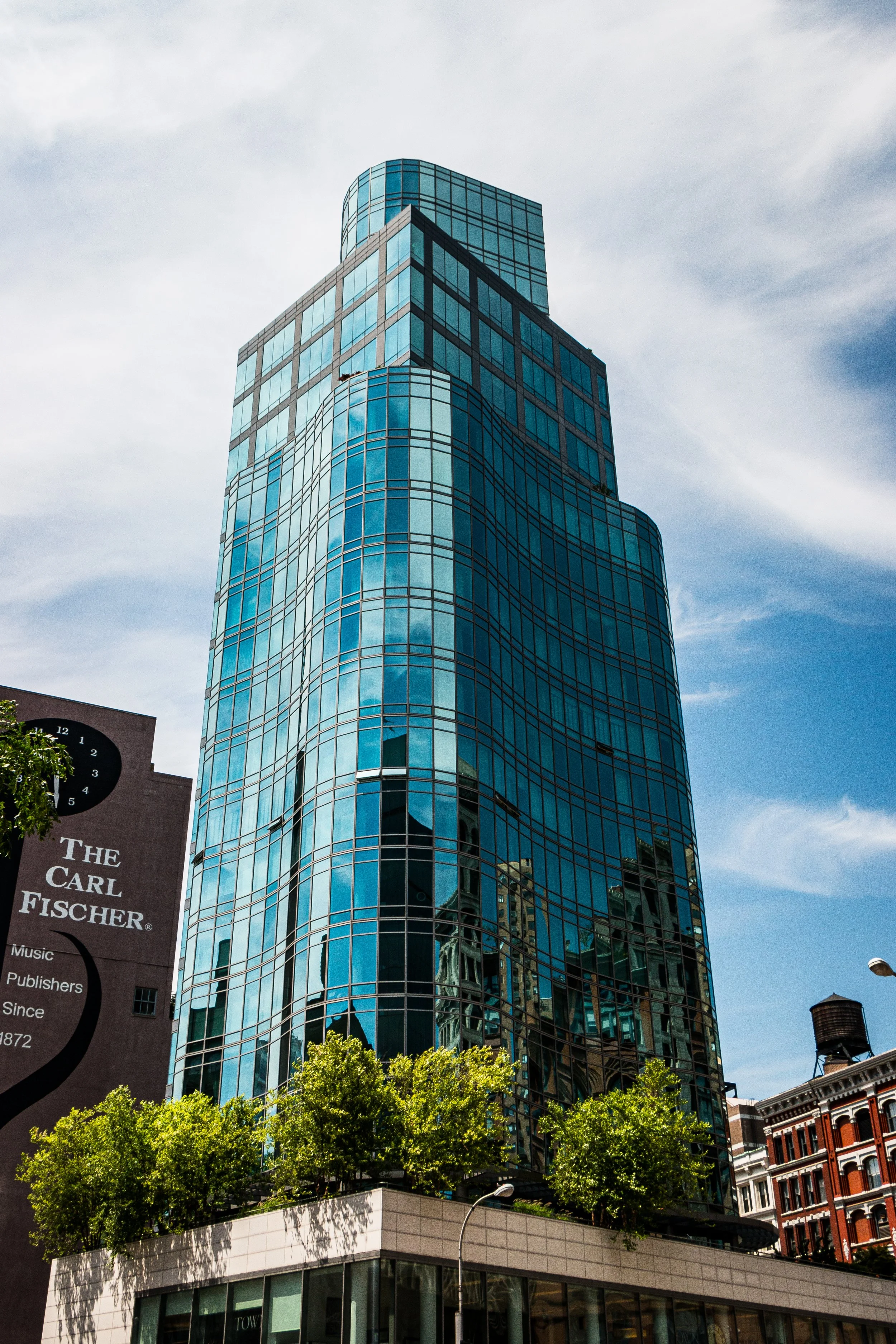 A tall modern glass skyscraper with reflective blue windows, situated in an urban area with nearby older buildings and green trees at the base.