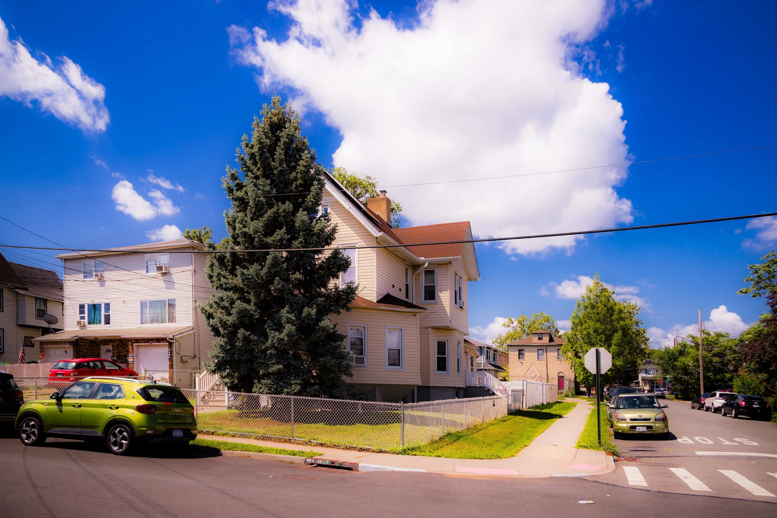 A residential street with a large yellow house, a tall evergreen tree, parked cars, a sidewalk, and a clear blue sky with scattered clouds.