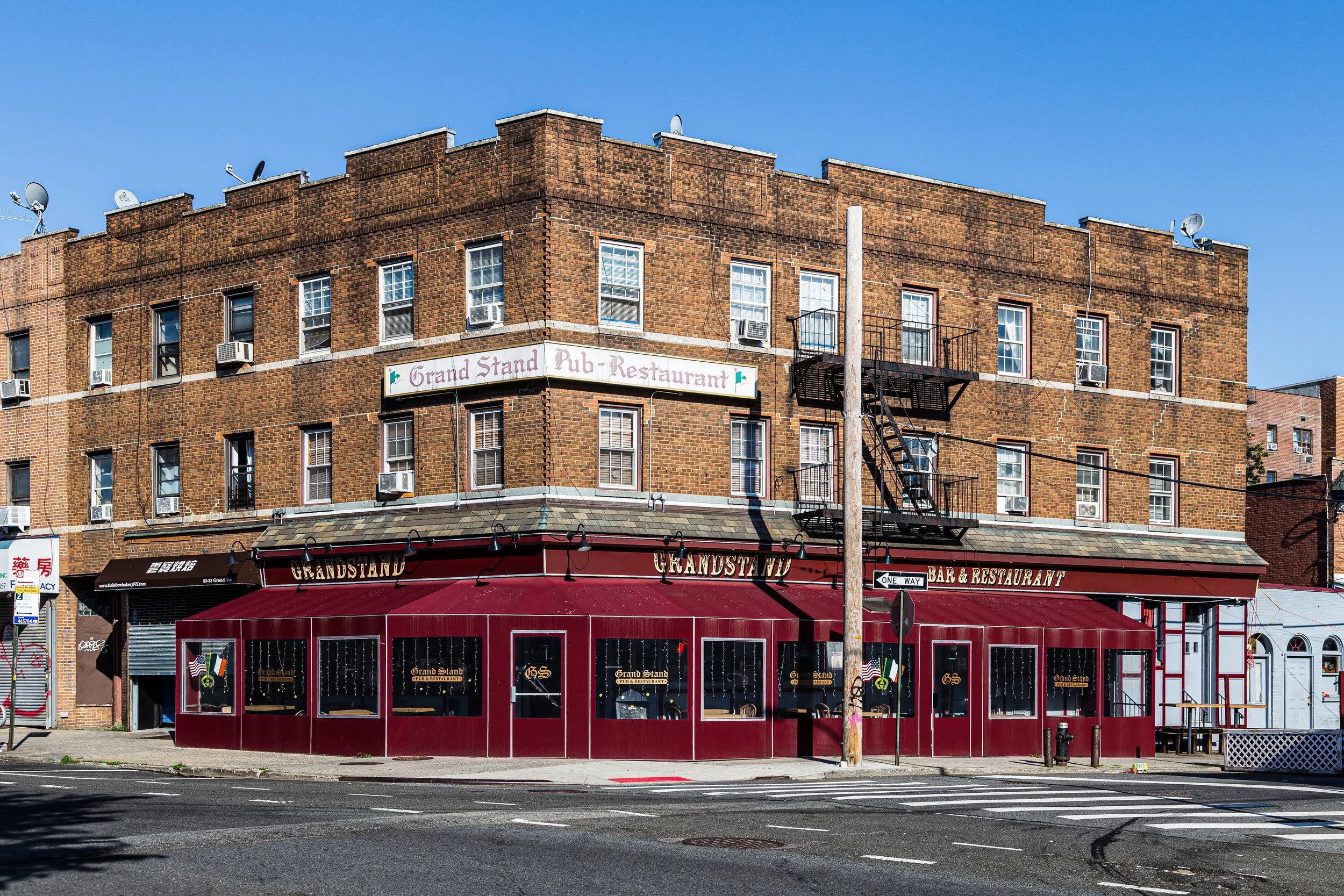 A multi-story brick building with a pub and restaurant on the ground floor. The restaurant has a red exterior with large windows and an awning, with signs reading 'Grandstand' and 'Bar & Restaurant.' The upper floors have multiple windows with air conditioning units, a fire escape, and a sign that reads 'Grand Stand Pub-Restaurant.' The street in front has a crosswalk, a fire hydrant, and a traffic sign, with a clear blue sky overhead.