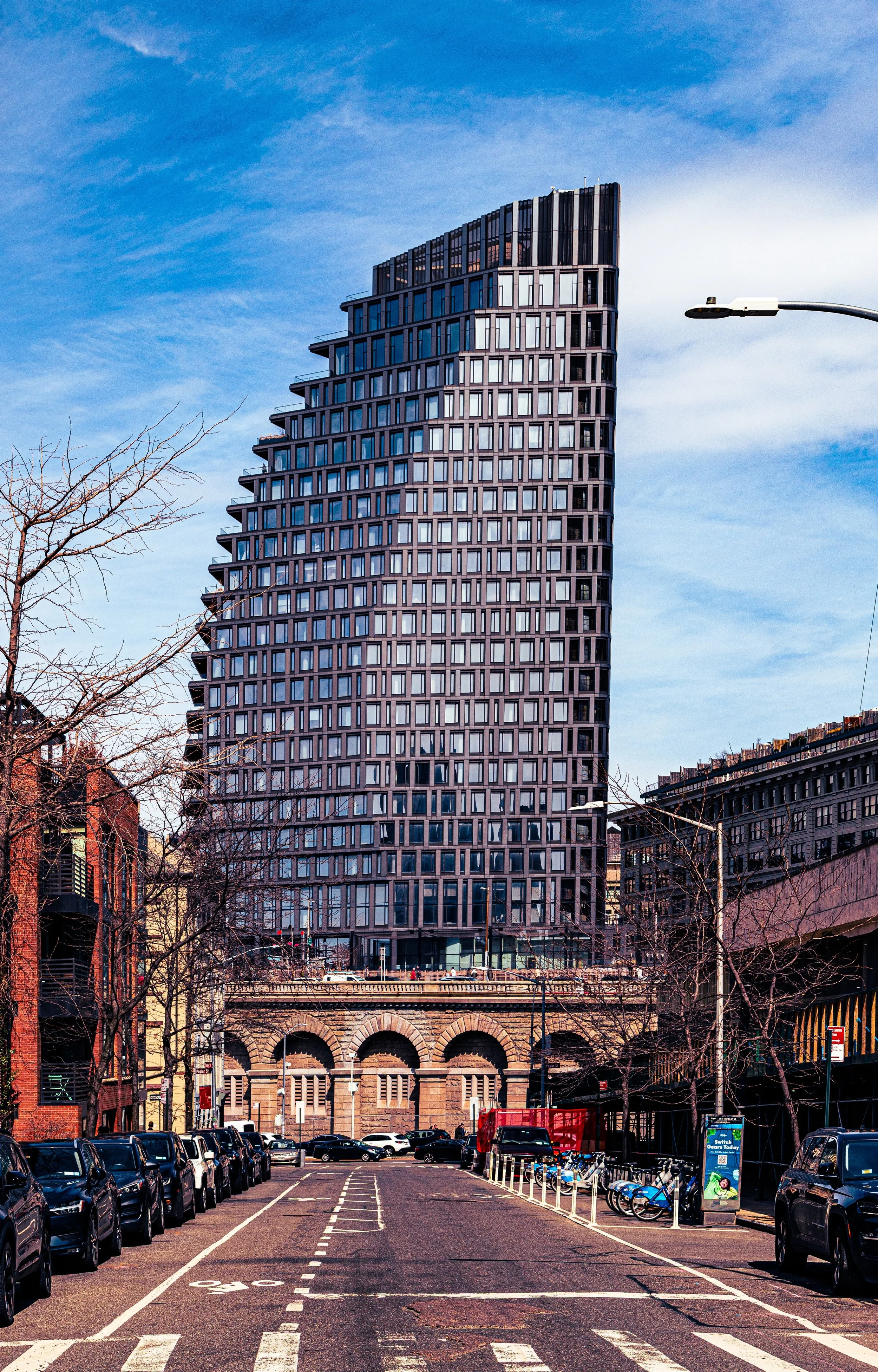 A tall modern high-rise building with numerous windows, set against a partly cloudy blue sky, with parked cars and trees in the foreground.