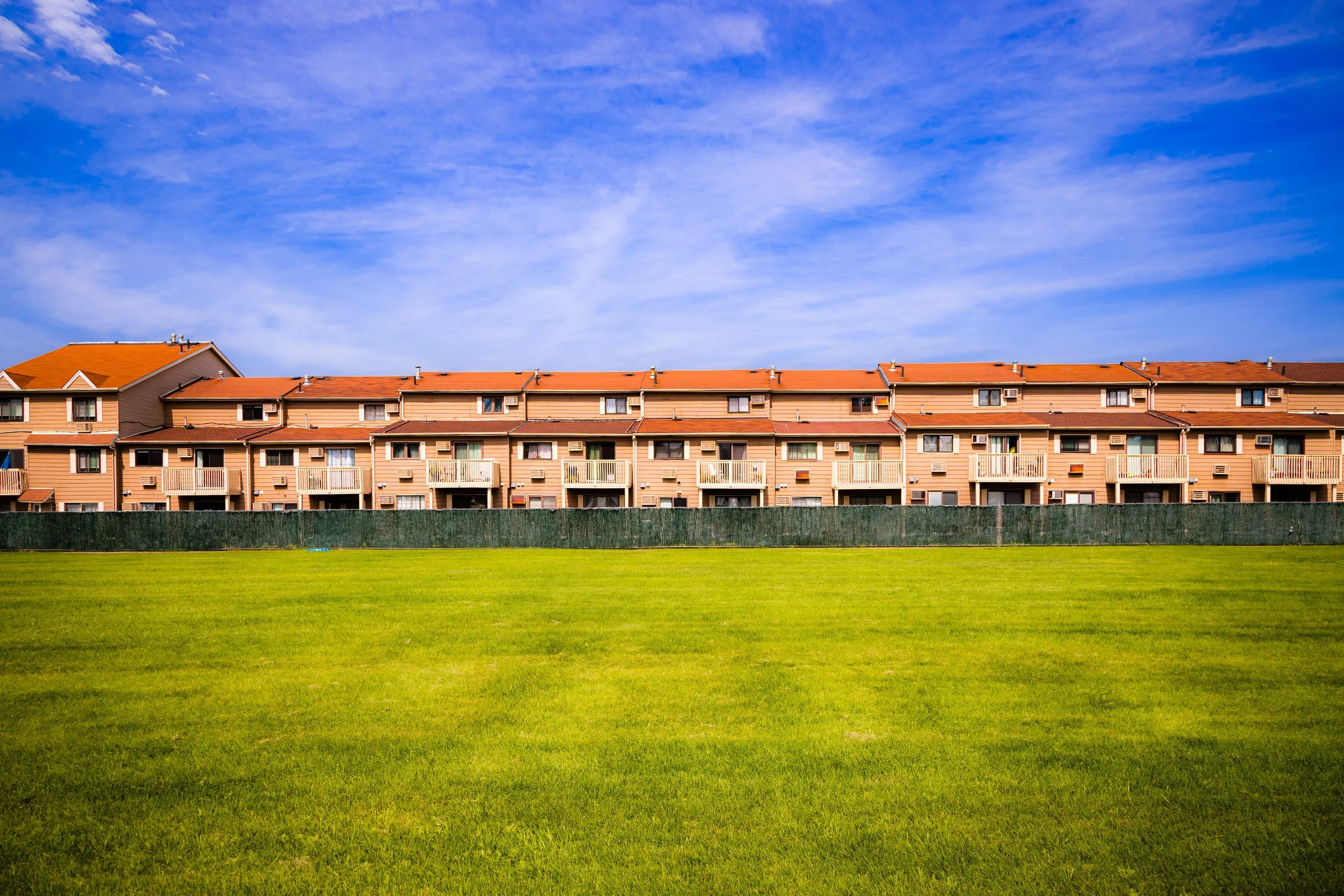 Residential apartment complex with multiple levels of balconies, set behind a green grassy field with a blue sky above.