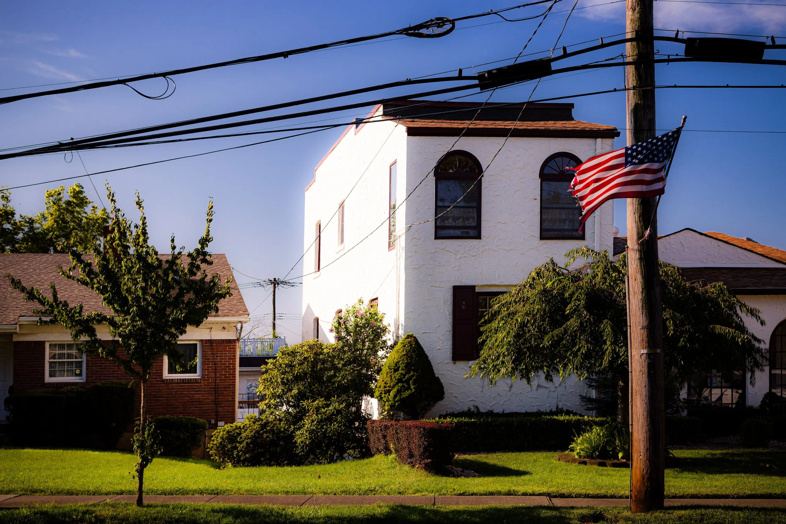 A residential street corner with a white stucco house, American flag on a wooden pole, and a small tree in the foreground. Power lines cross the blue sky.