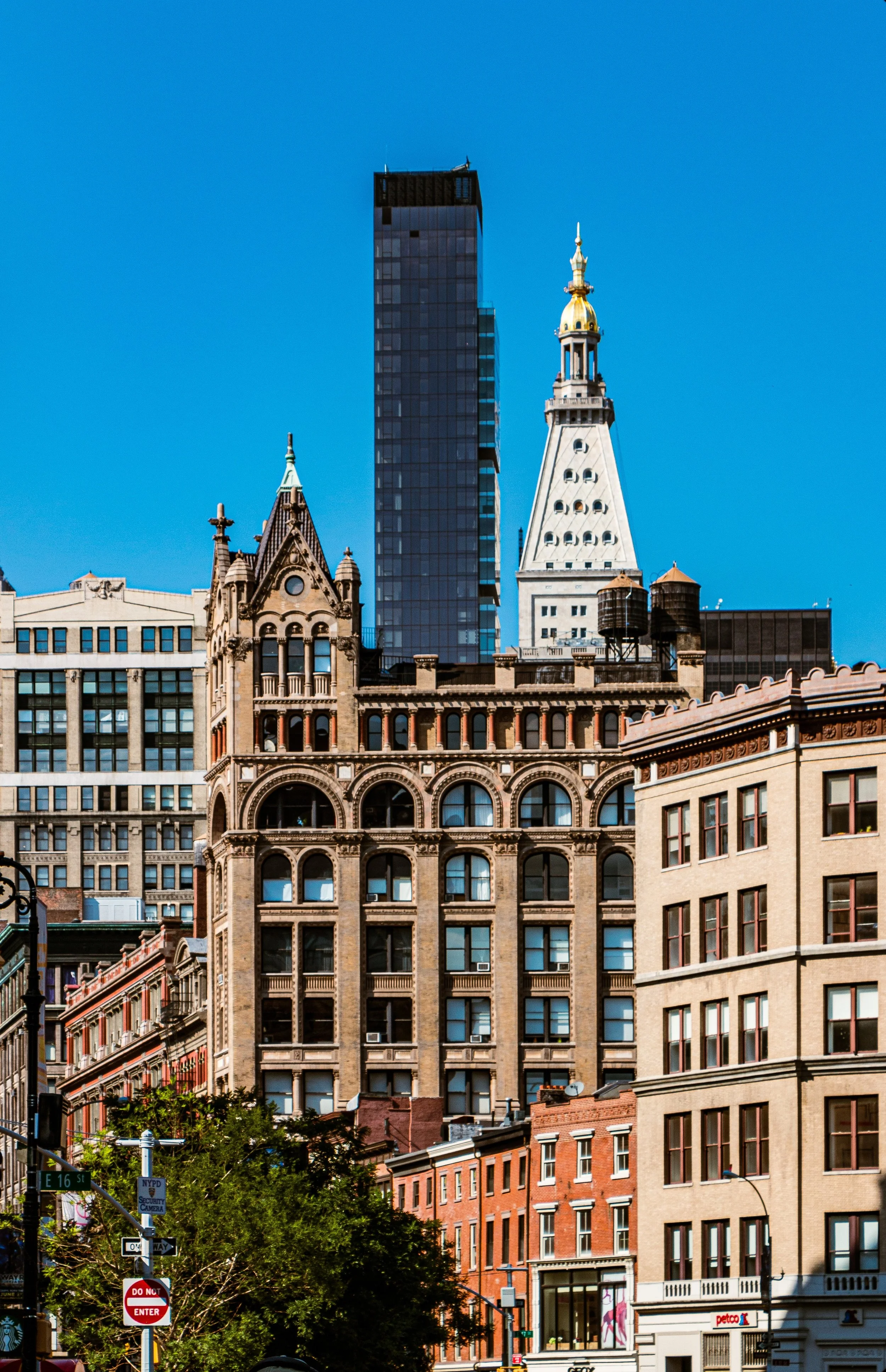 Cityscape featuring historic and modern buildings under a clear blue sky, with trees and street signs in the foreground.