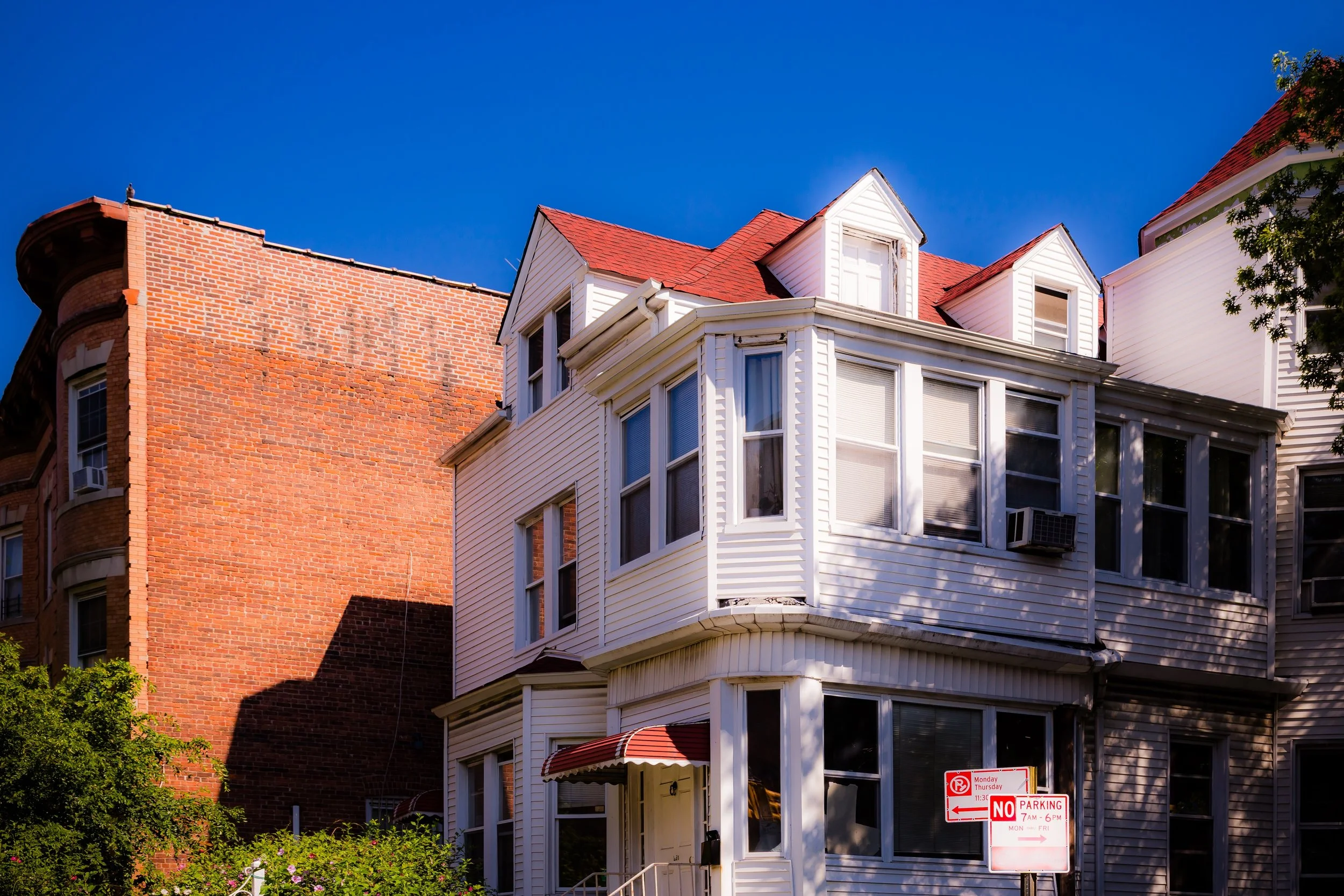 A white multi-story house with bay windows and a red roof, adjacent to a tall brick building, under a bright blue sky.