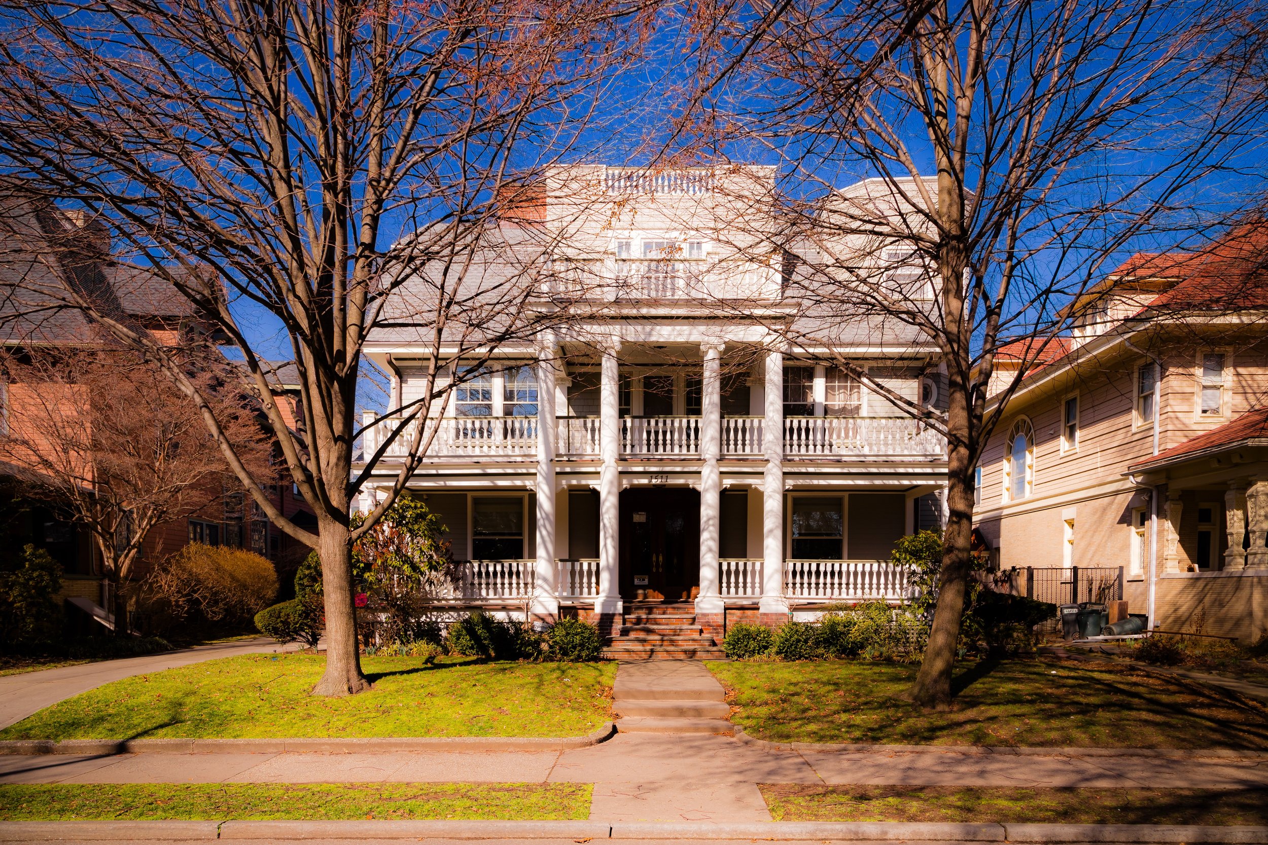 A large, white, three-story house with a porch and columns, trees with no leaves, and a well-maintained front yard with grass and bushes, under a clear blue sky.