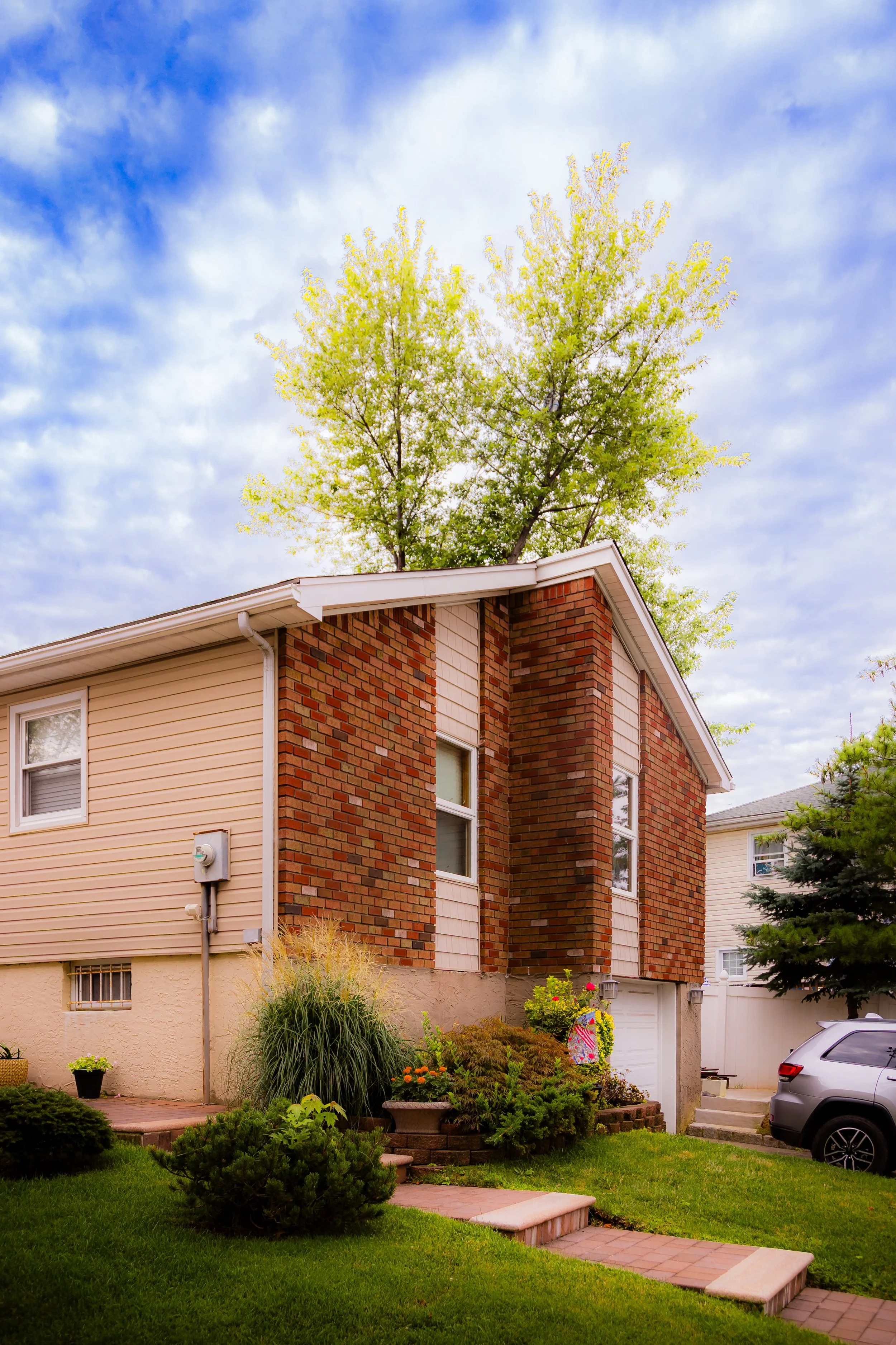 A two-story house with beige siding and brick exterior, surrounded by a green lawn and plants, under a partly cloudy sky.
