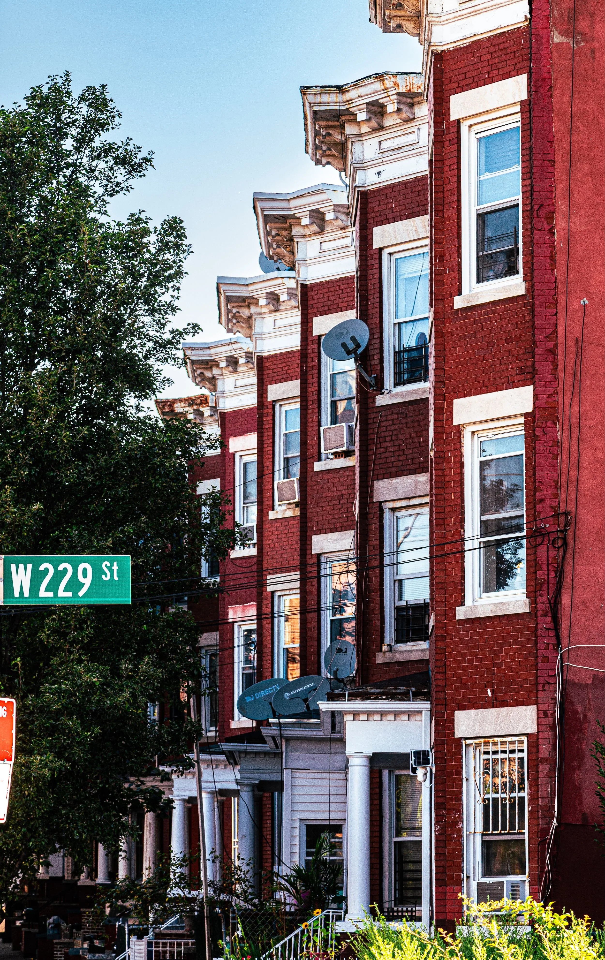 Red brick apartment building with white decorative trim, multiple satellite dishes on the exterior, and a street sign showing W 229th Street. The building has several windows, some with air conditioning units, and a small front yard with plants and a white fence.