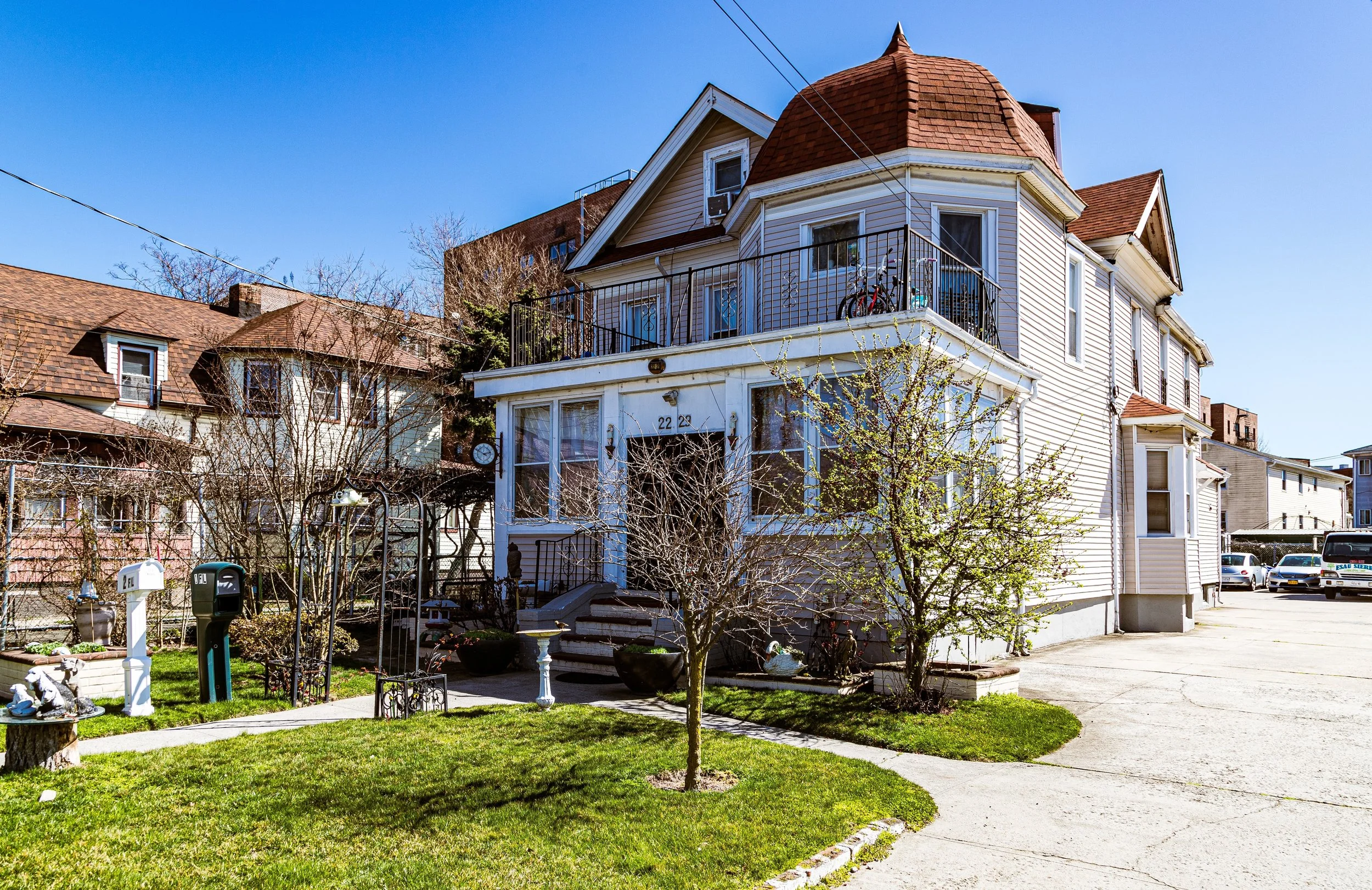 A two-story Victorian house with a rounded turret and a front porch, surrounded by small trees, a well-maintained lawn, and a suburban street with parked cars.