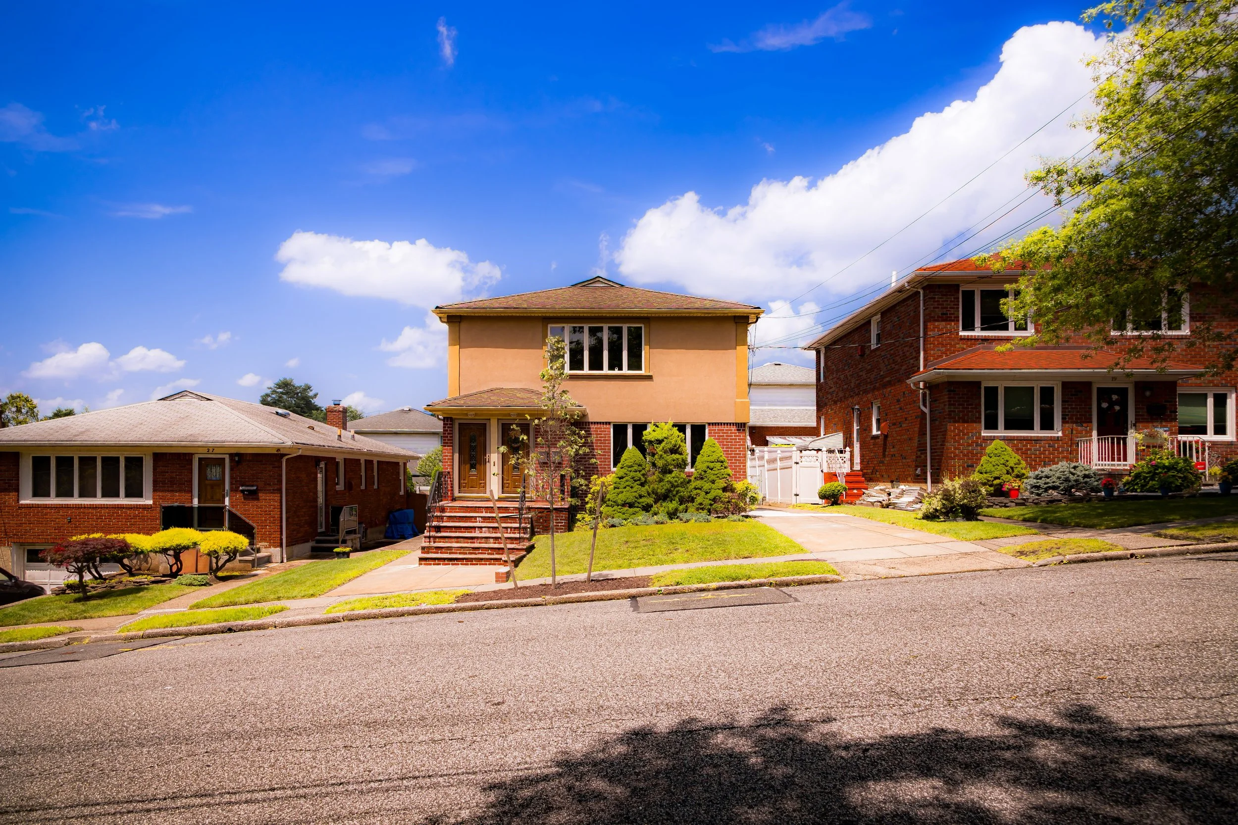 A residential neighborhood with three houses, including a central two-story house with beige and brick exterior, front yard with small trees and bushes, and a clear blue sky with some clouds.