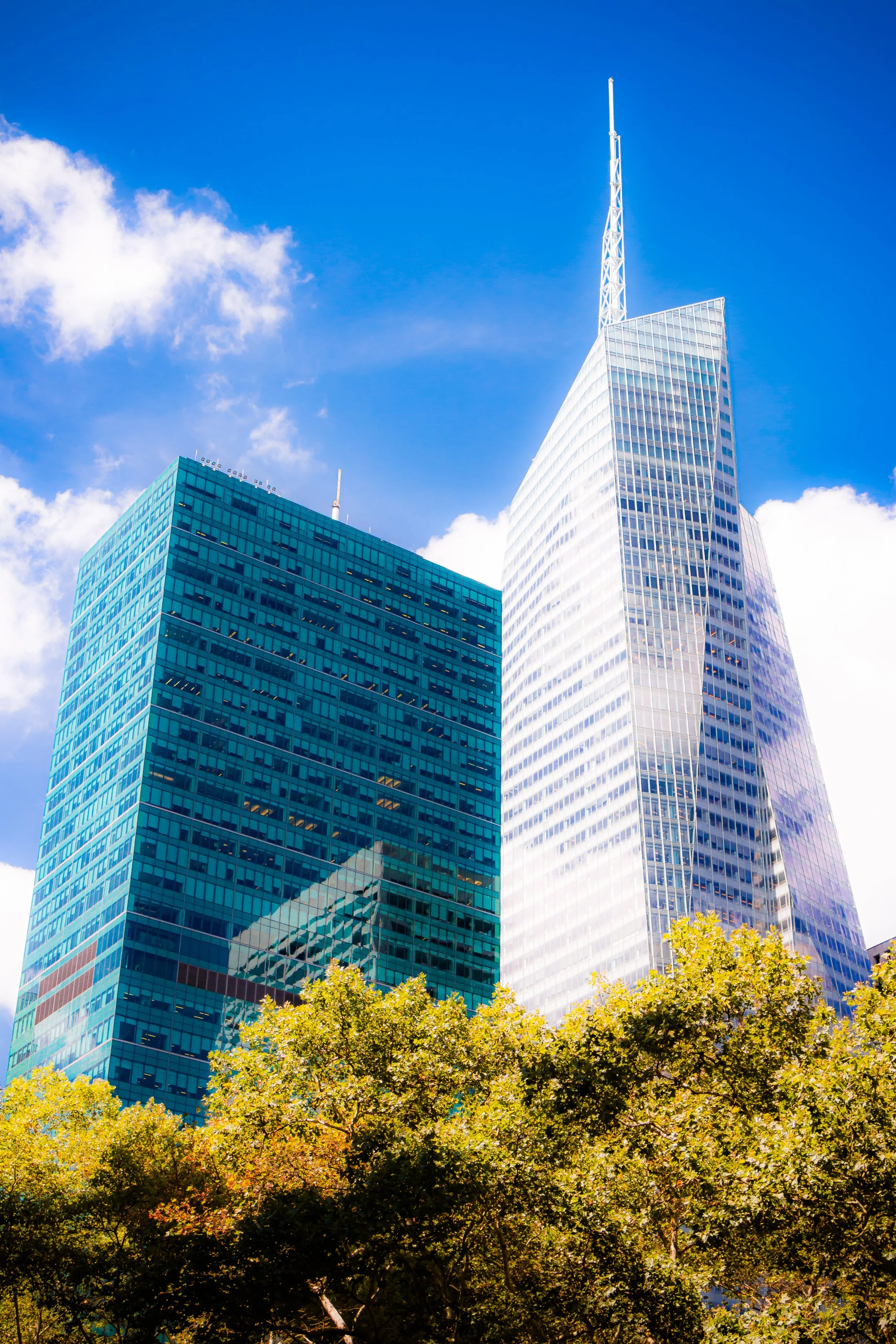 Two modern skyscrapers with glass facades, one with a pointed spire, against a blue sky with some clouds, and trees with green foliage at the base.