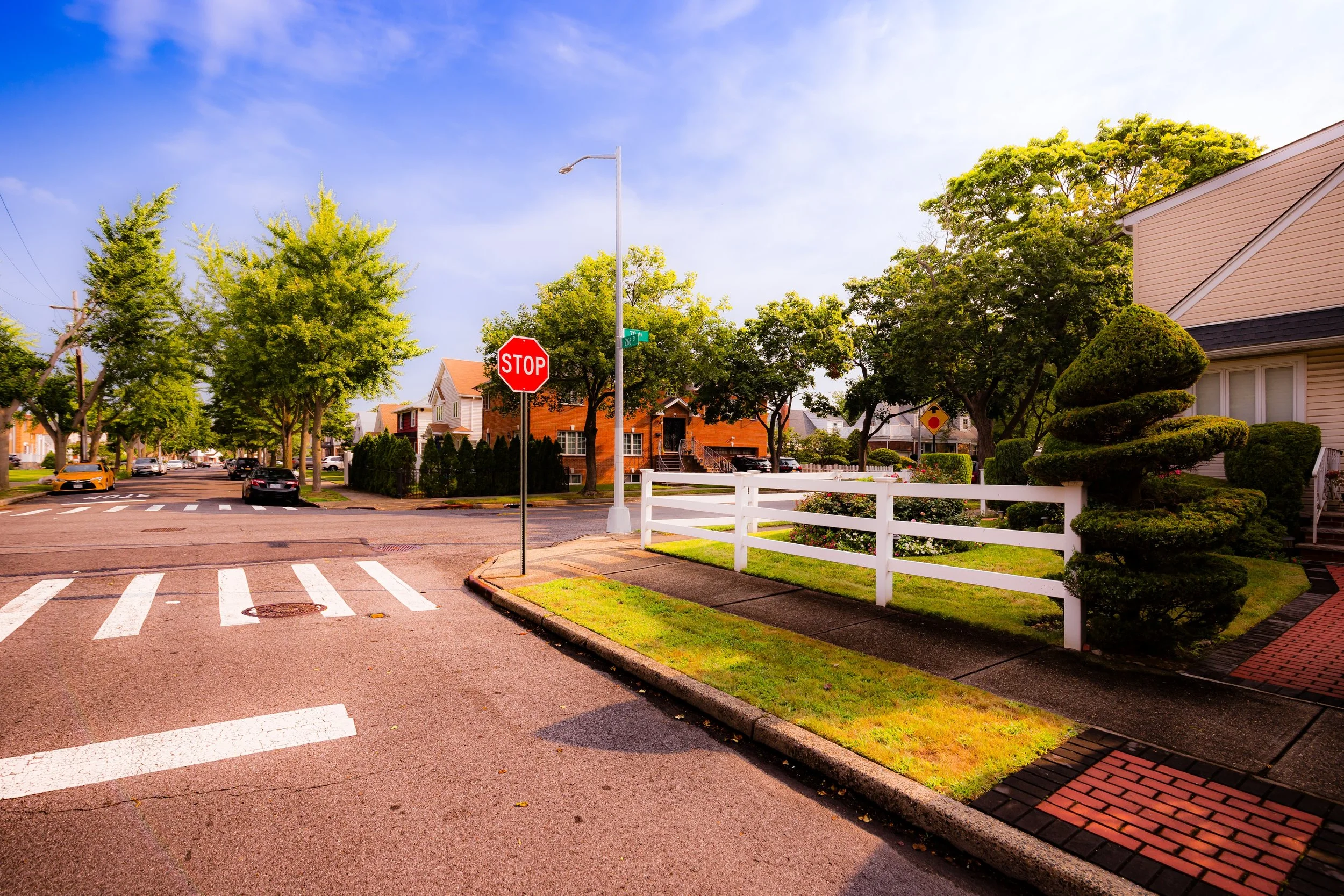 A sunny suburban street with a stop sign at the crosswalk, lush green trees, a well-maintained yard with shaped bushes, and residential houses in the background.