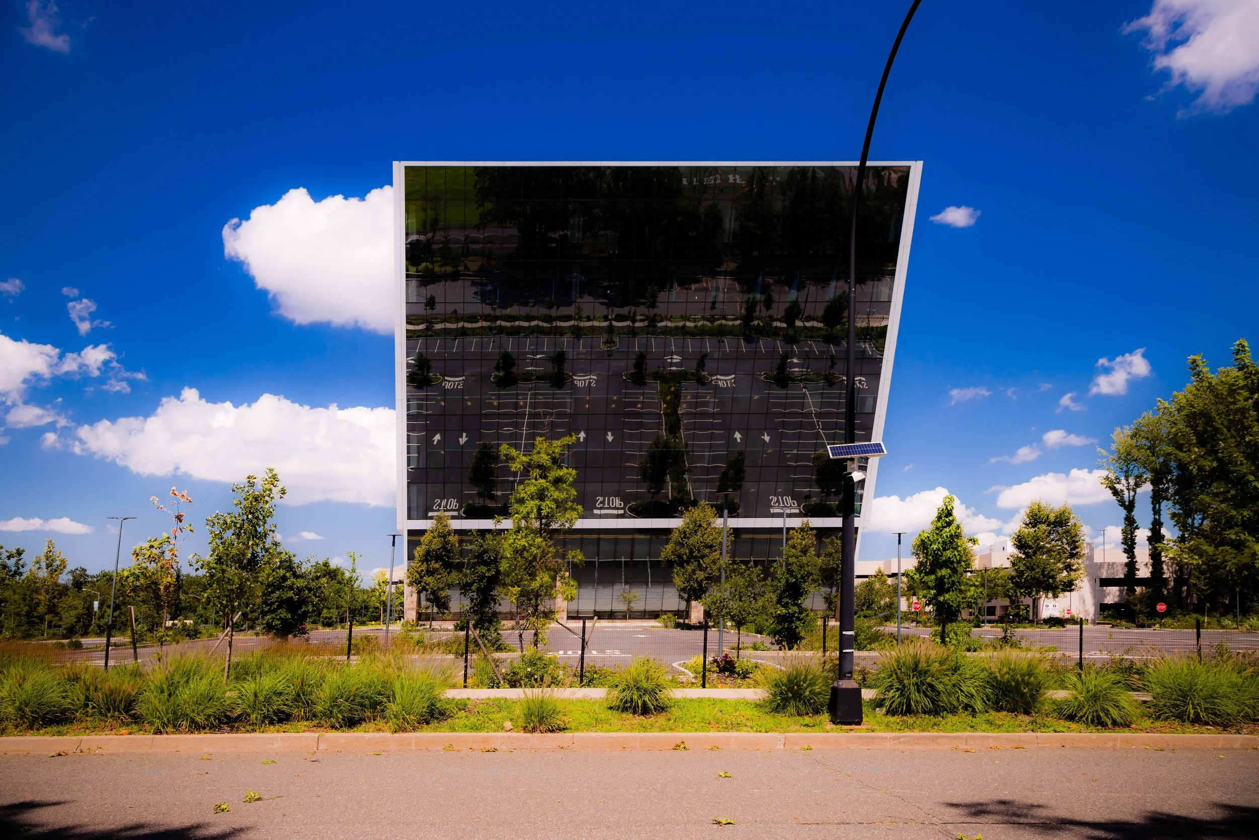 Modern glass building with reflective windows, surrounded by trees, sidewalk, and parking lot under a blue sky with clouds.