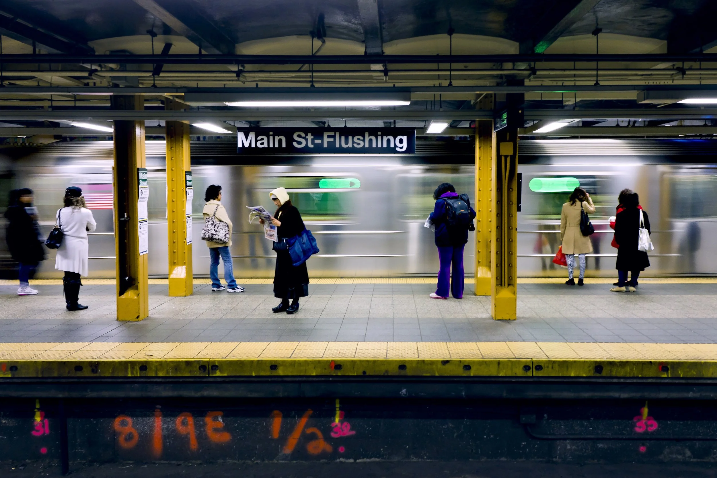 People waiting on a subway platform at Main St-Flushing station with a train passing by.