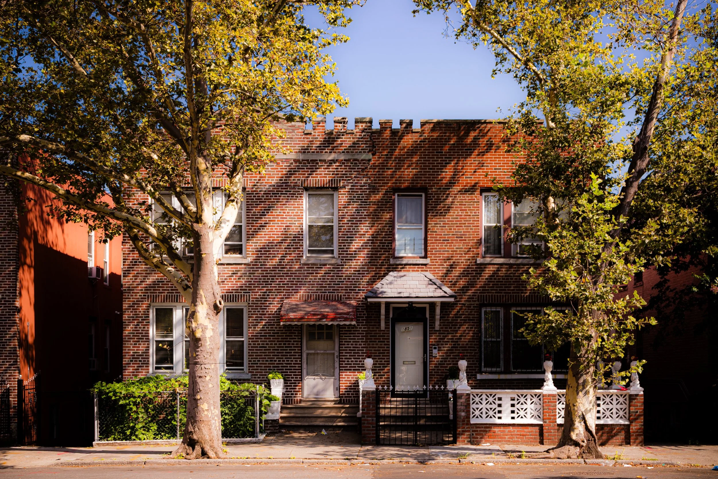 A brick townhouse with three stories, a small front porch, two trees in front, and a clear blue sky.