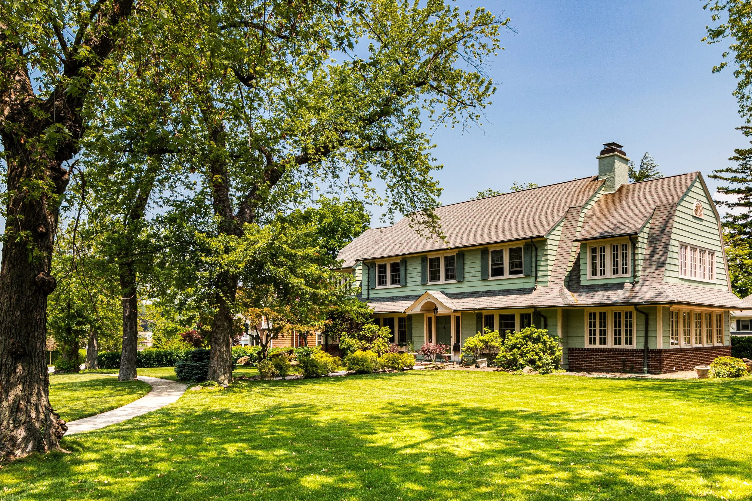 A large green house with yellow shutters and brick foundation surrounded by a well-maintained garden and large trees, under a clear blue sky.