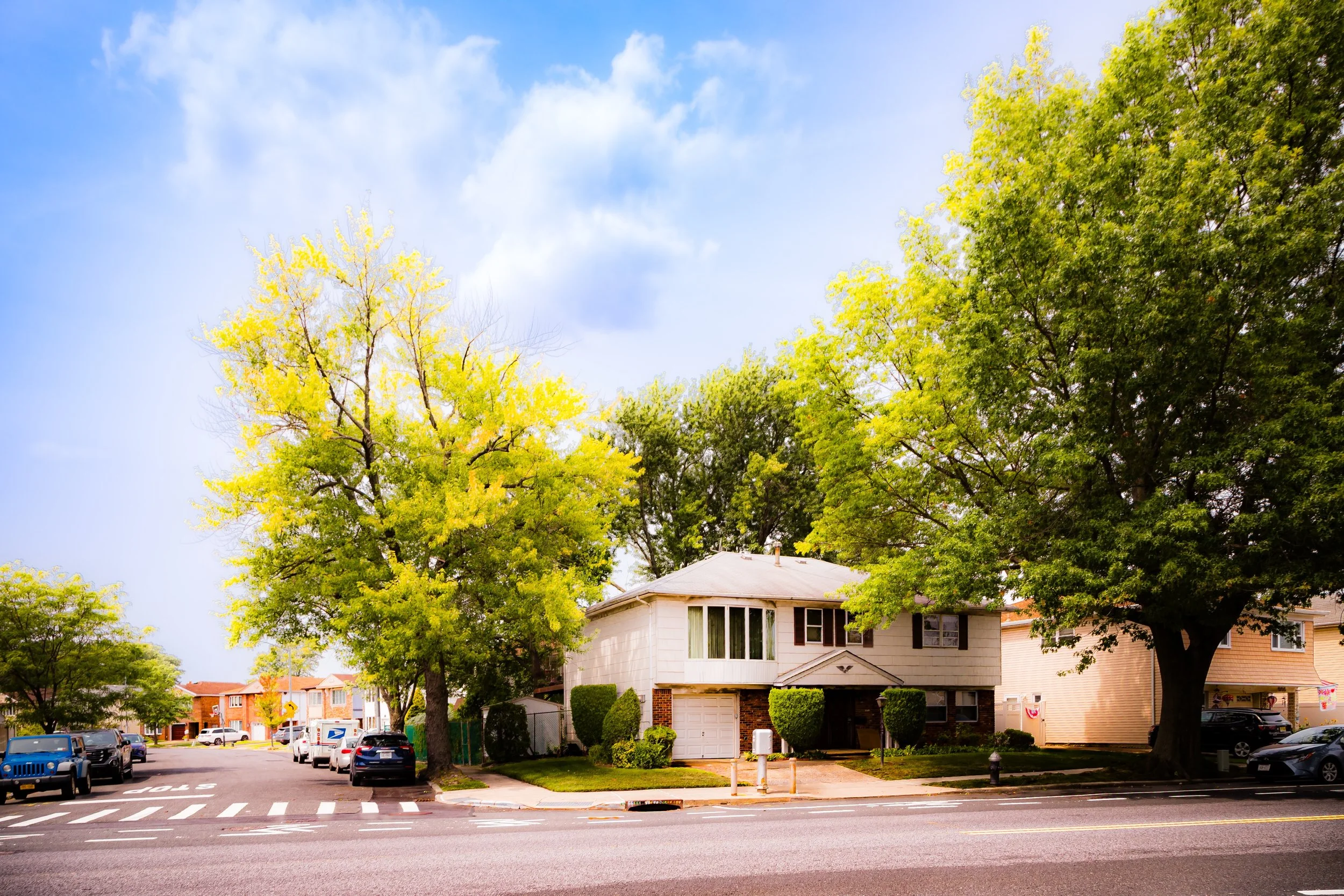 A residential street with a house, large trees, parked cars, and a sidewalk under a partly cloudy sky.